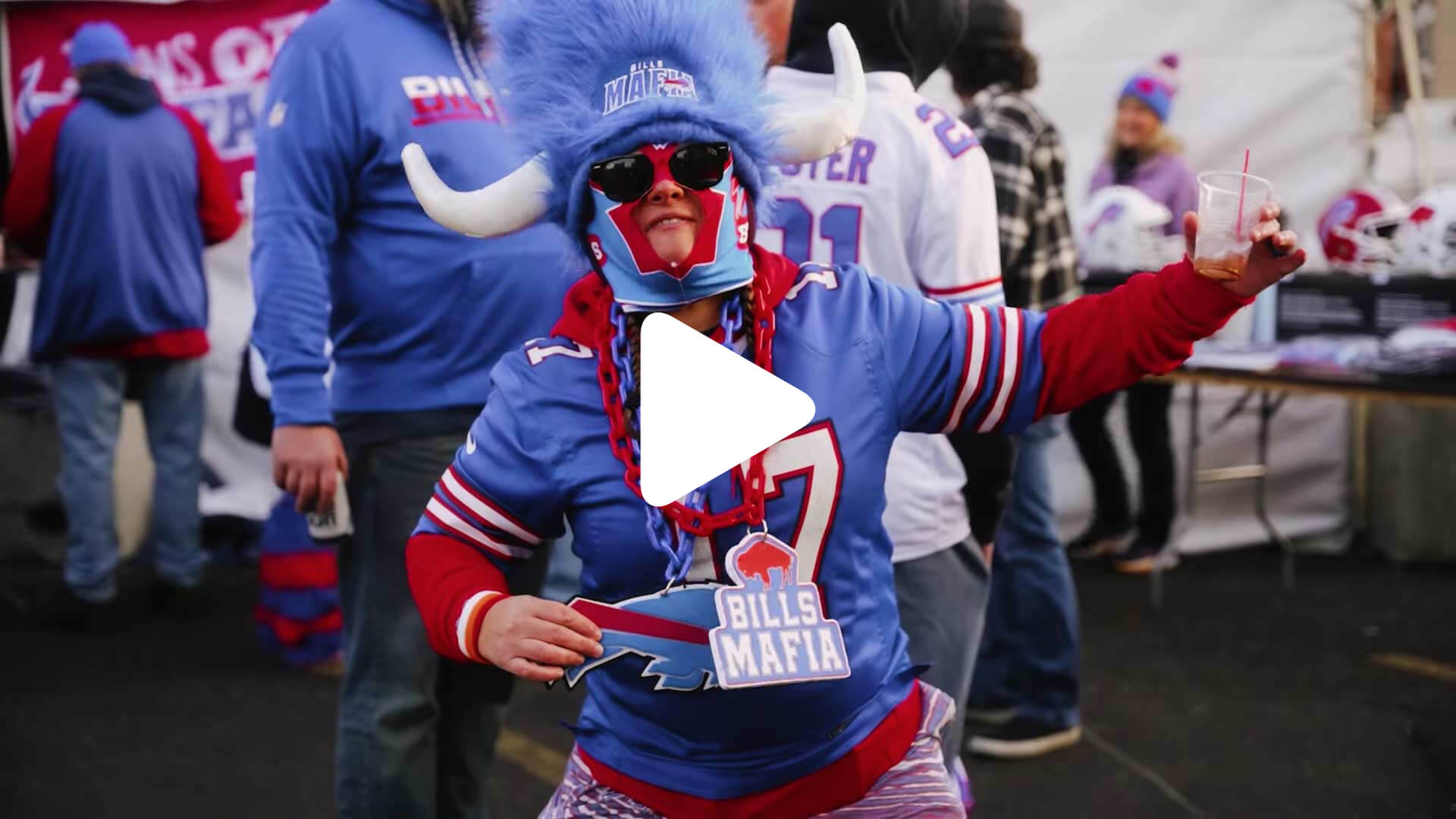 A person wearing a blue buffalo hat, sunglasses, and a Buffalo Bills jersey poses energetically at an outdoor event, holding a "Bills Mafia" sign. Other people in Bills gear are visible in the background.