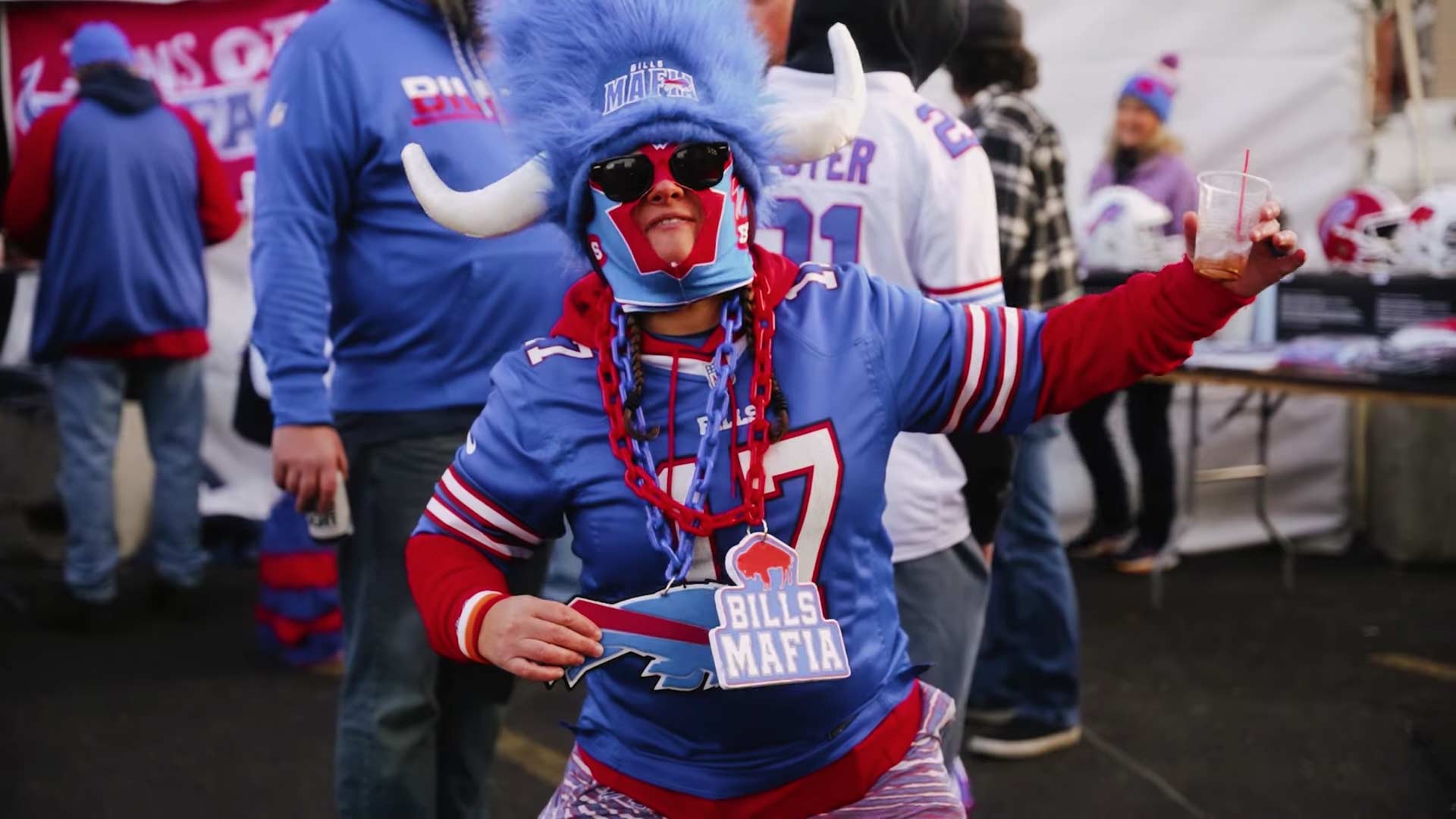 A fan wearing a large blue furry hat with horns, sunglasses, and a “Bills Mafia” necklace poses energetically at an outdoor event, dressed in Buffalo Bills gear among other fans.