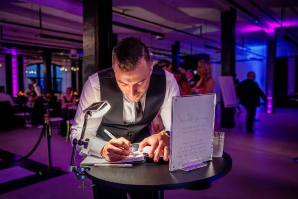 A man in formal attire leans over a small round table, writing on paper with a pen. The table holds a glass of water and a display sign. The background shows people and purple event lighting.