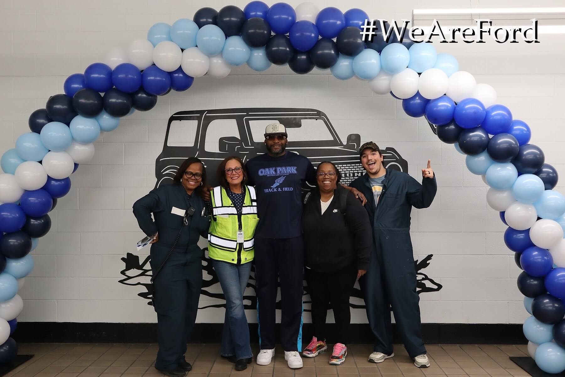 Five people stand together and smile under a blue and white balloon arch in front of a wall with a car illustration. Four wear work uniforms; one wears a reflective vest. The text "#WeAreFord" appears in the top right corner.