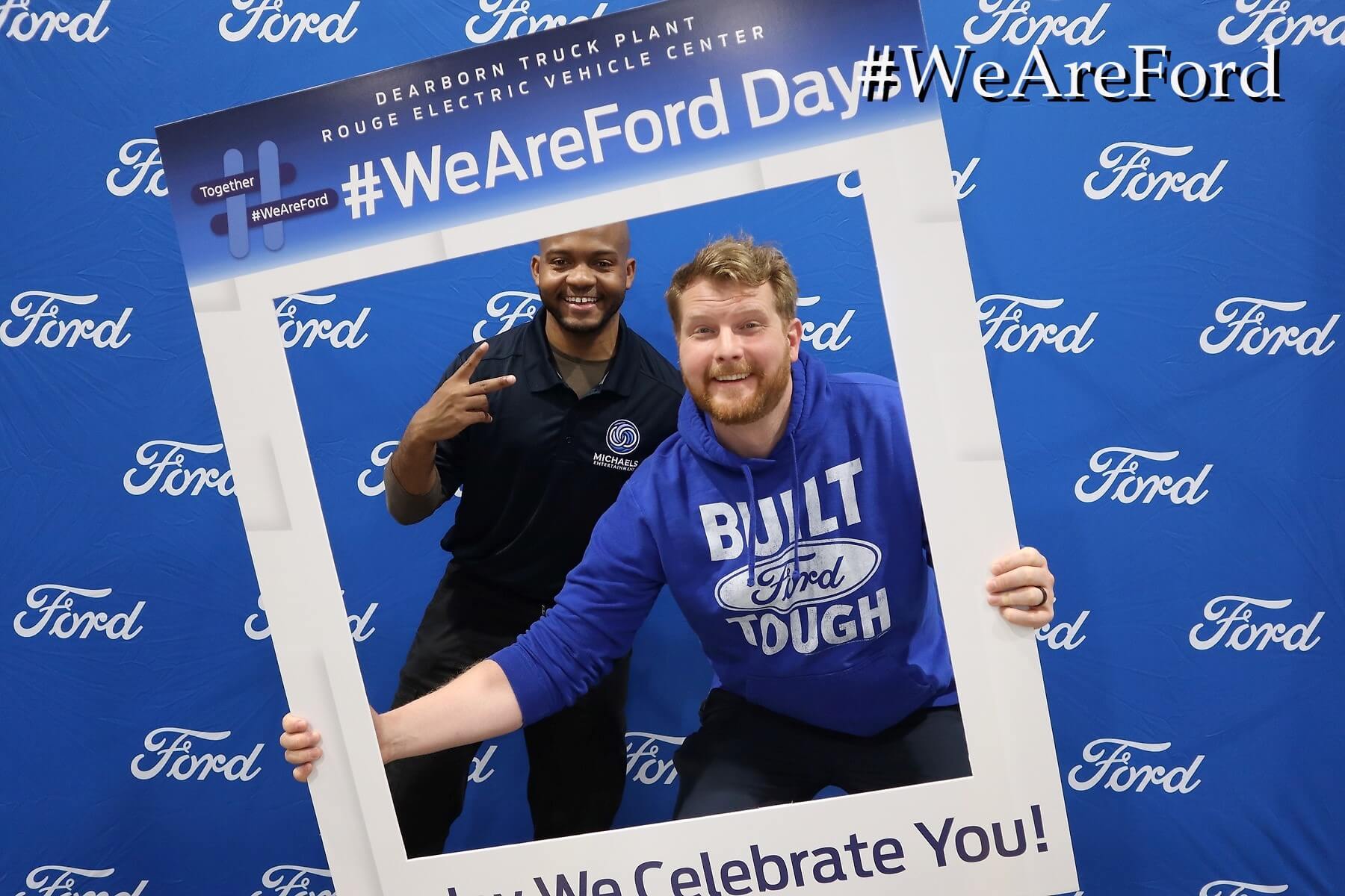 Two men pose and smile behind a large photo frame cutout with "#WeAreFord Day" printed on it, in front of a blue Ford-branded backdrop. One man wears a "Built Ford Tough" hoodie; the other flashes a peace sign.