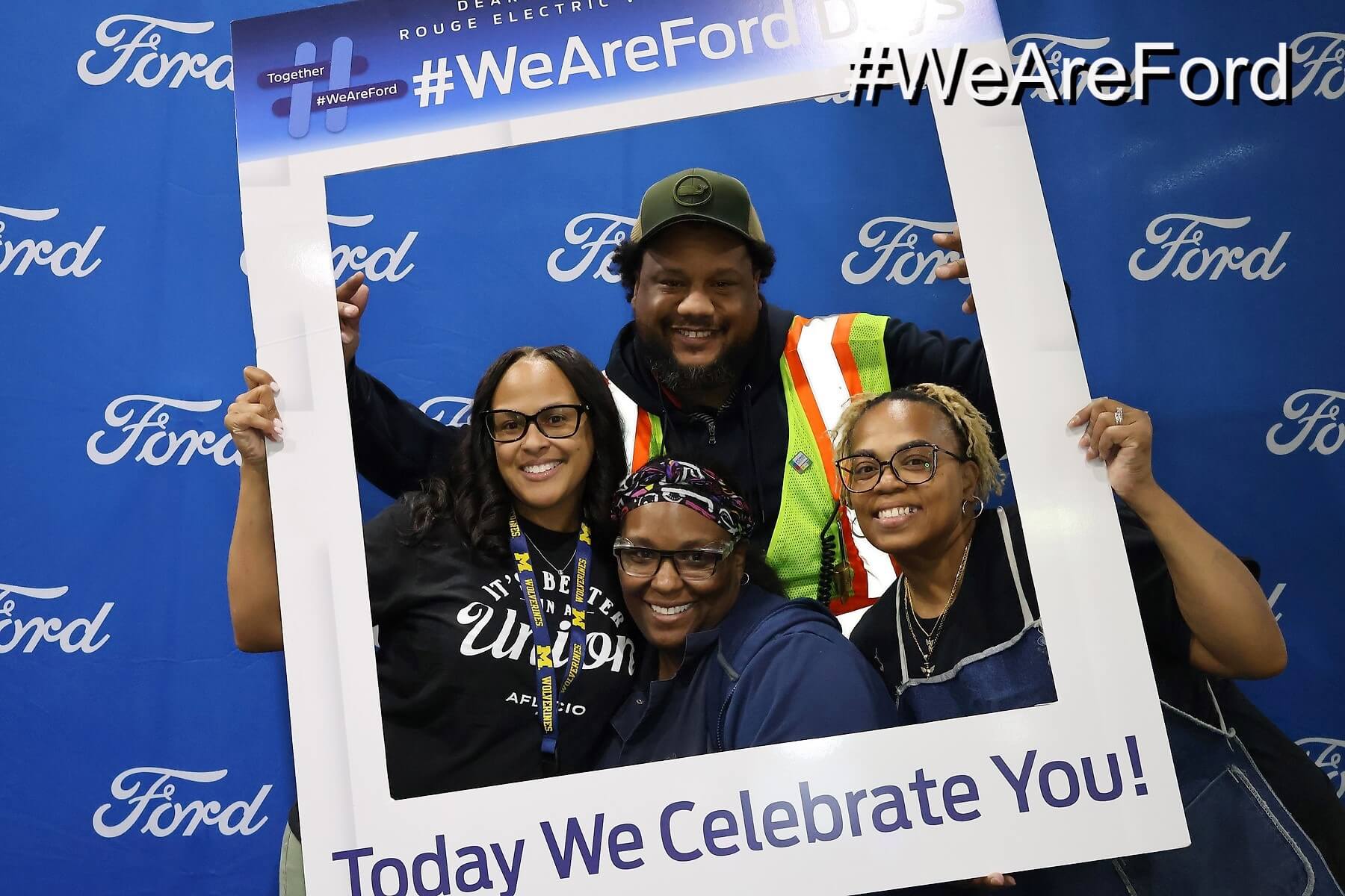 Four people pose and smile behind a large photo frame with the text “Today We Celebrate You!” in front of a blue backdrop with repeated Ford logos and #WeAreFord signage.