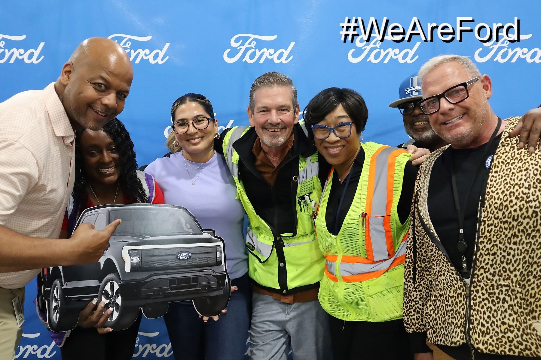 A group of seven people stand in front of a blue Ford backdrop, smiling. One person holds a cardboard cutout of a Ford truck. Two people wear safety vests. The hashtag "#WeAreFord" appears in the corner.