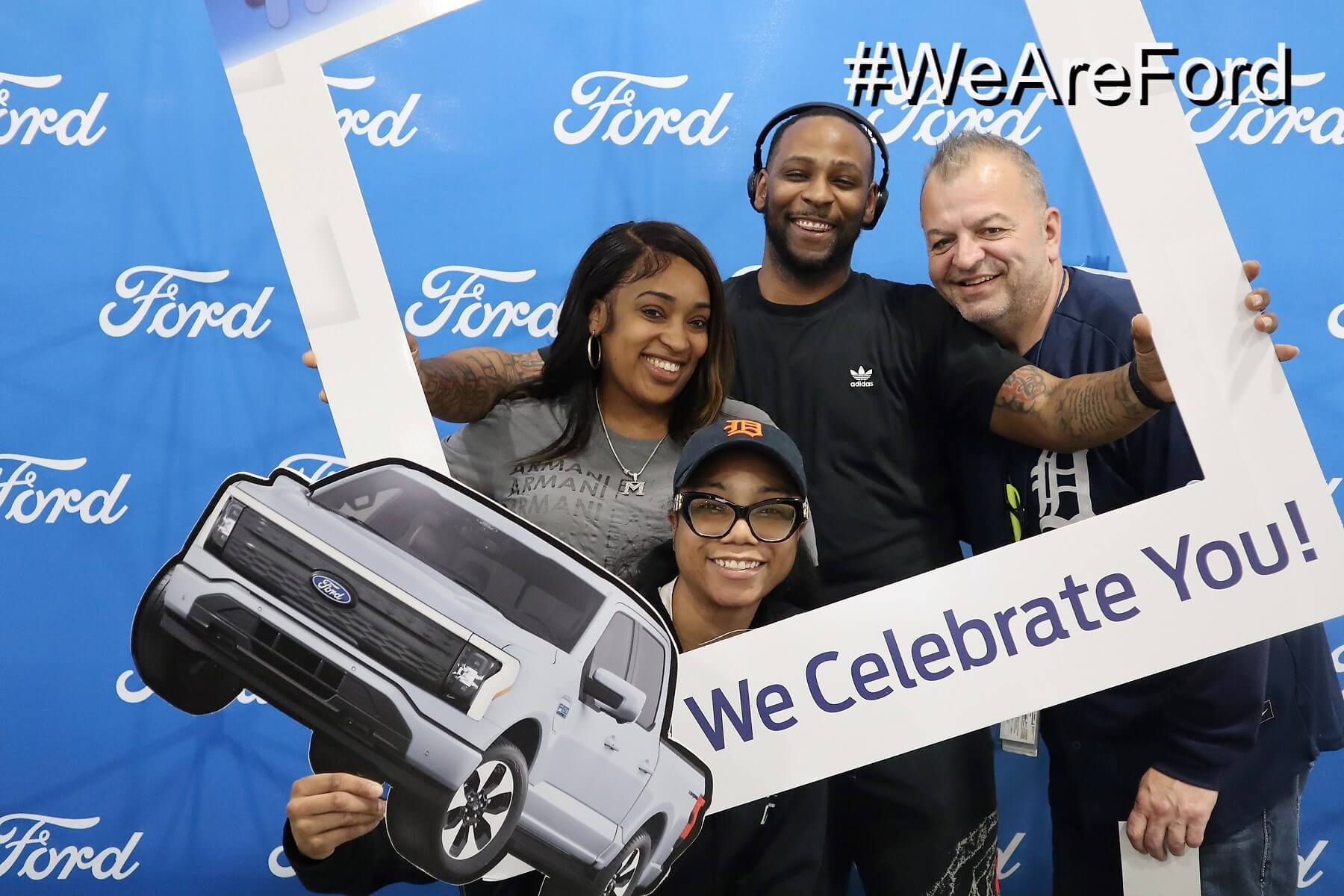 Four people smile while holding a large white frame and a cardboard cutout of a Ford truck in front of a blue Ford-branded backdrop. The frame has the words "We Celebrate You!" and the hashtag "#WeAreFord.