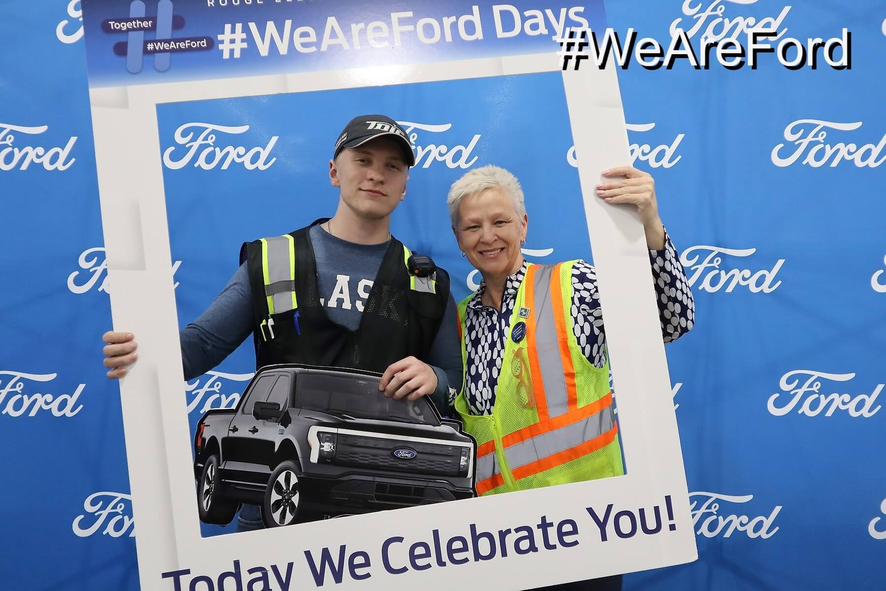 Two people pose and smile together, holding a large frame with an image of a Ford truck and the text "Today We Celebrate You!" in front of a blue Ford-branded backdrop. Hashtags #WeAreFord are visible.