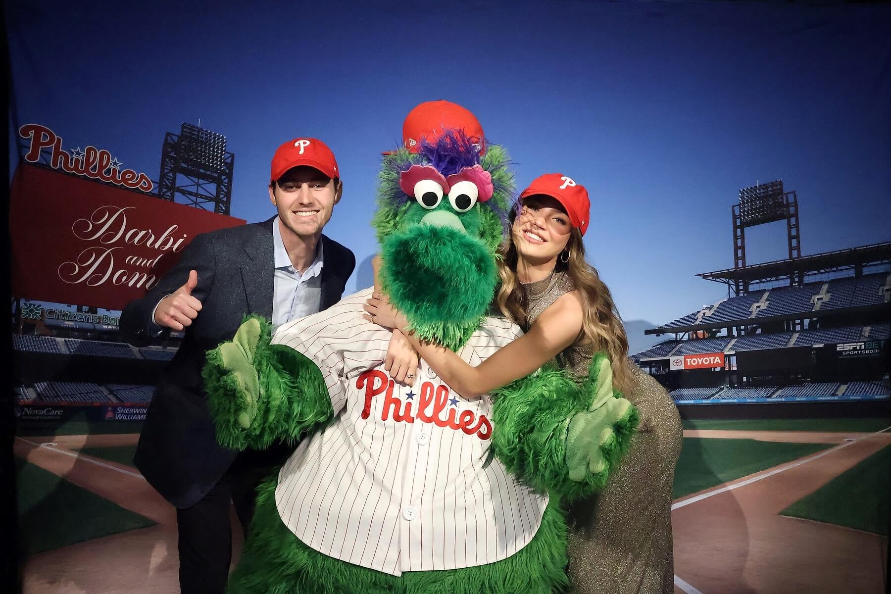 Two people wearing red Phillies caps pose and smile with the Phillie Phanatic mascot, who is dressed in a Phillies jersey and cap, on a baseball field with stadium seats and a scoreboard in the background.