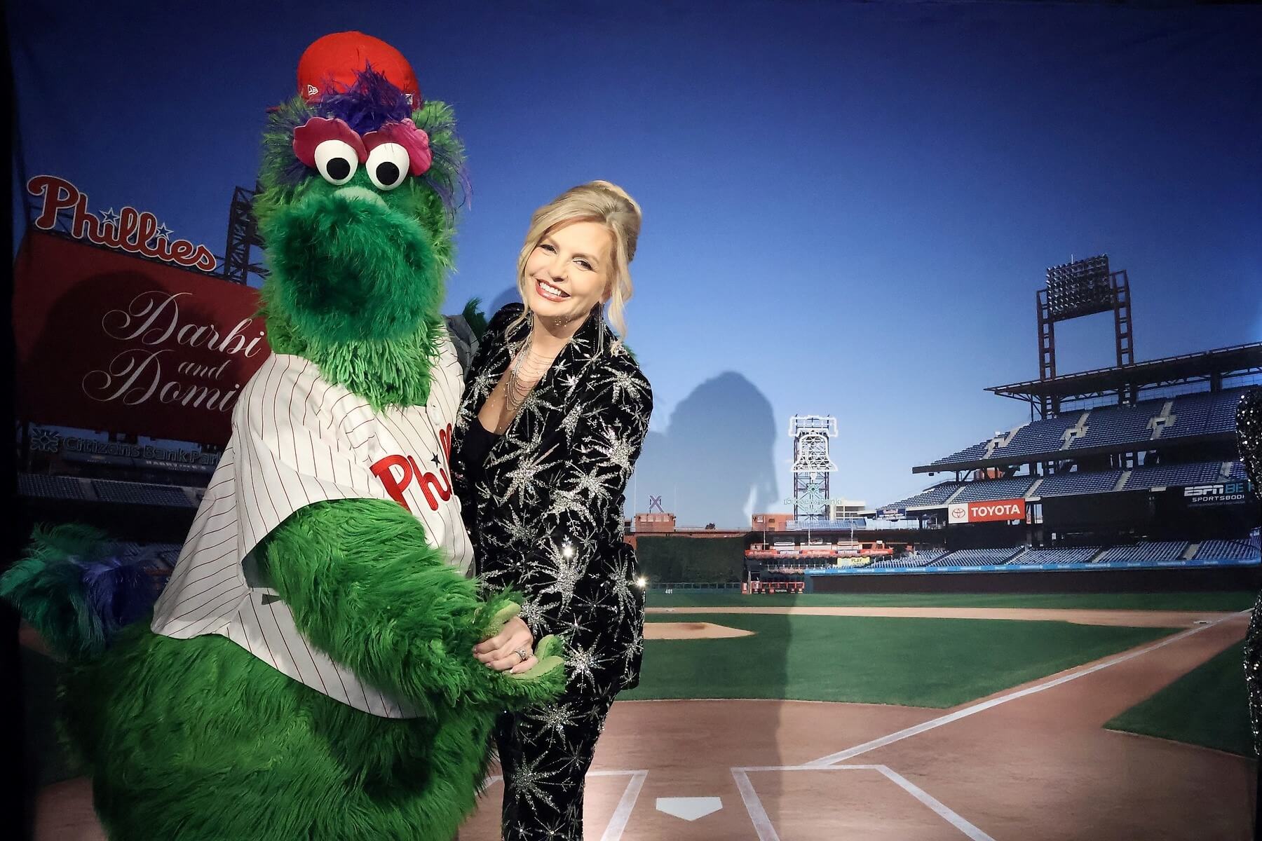 A woman in a black and silver outfit stands next to the Phillie Phanatic mascot on a baseball field backdrop, both smiling and holding hands near home plate.