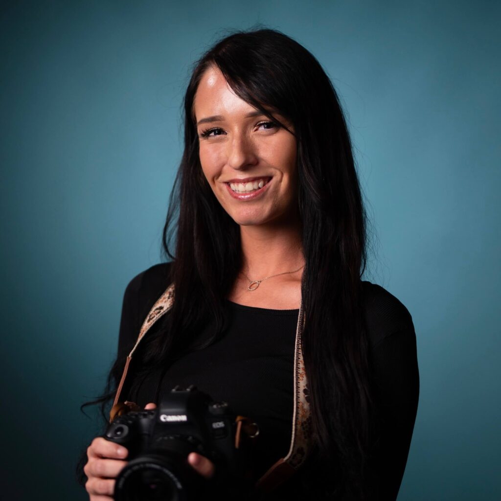 A woman with long dark hair smiles at the camera while holding a Canon camera against a plain blue background. She is wearing a black top and a camera strap around her neck.