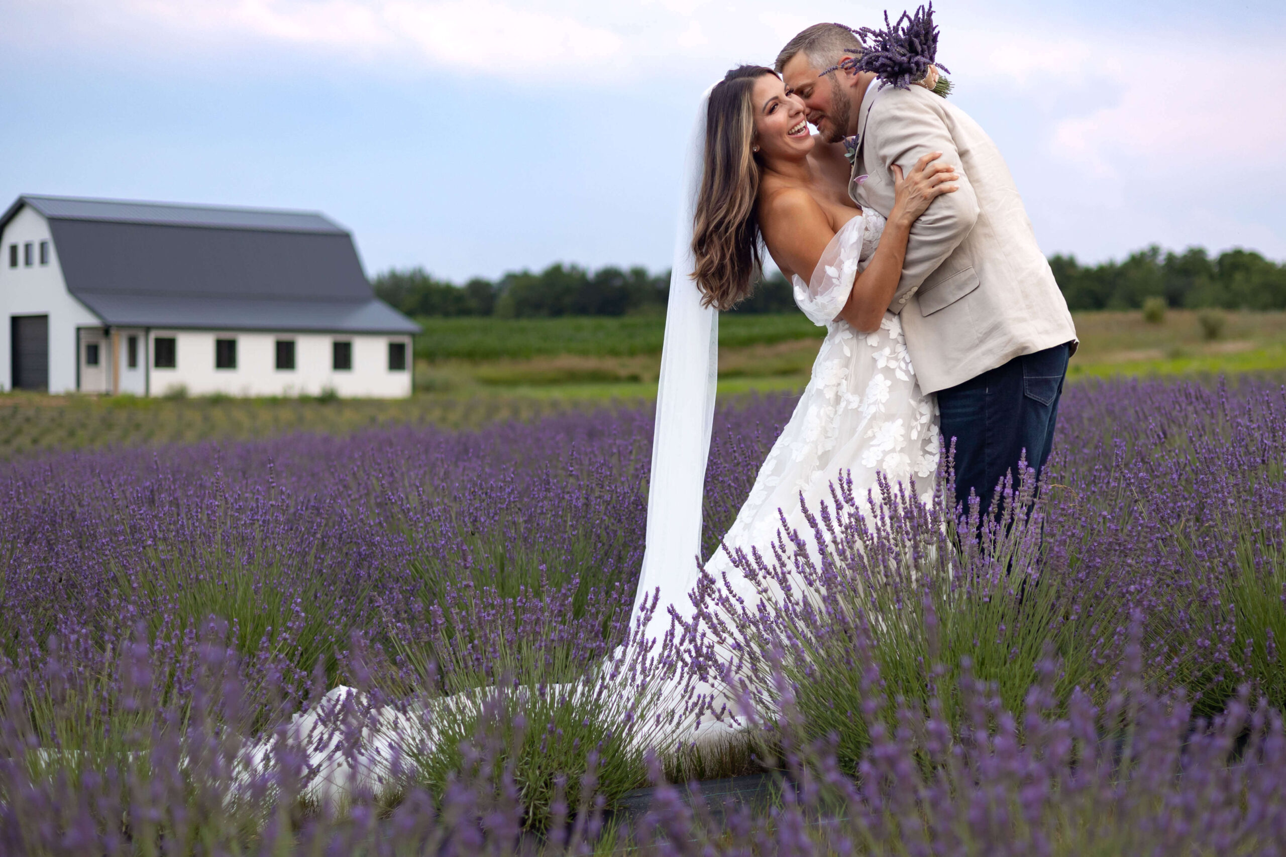 A bride and groom embrace and smile in a field of blooming lavender, with a white barn in the background and green trees under a cloudy sky. The bride wears a white dress and veil, and the groom holds a bouquet of lavender.