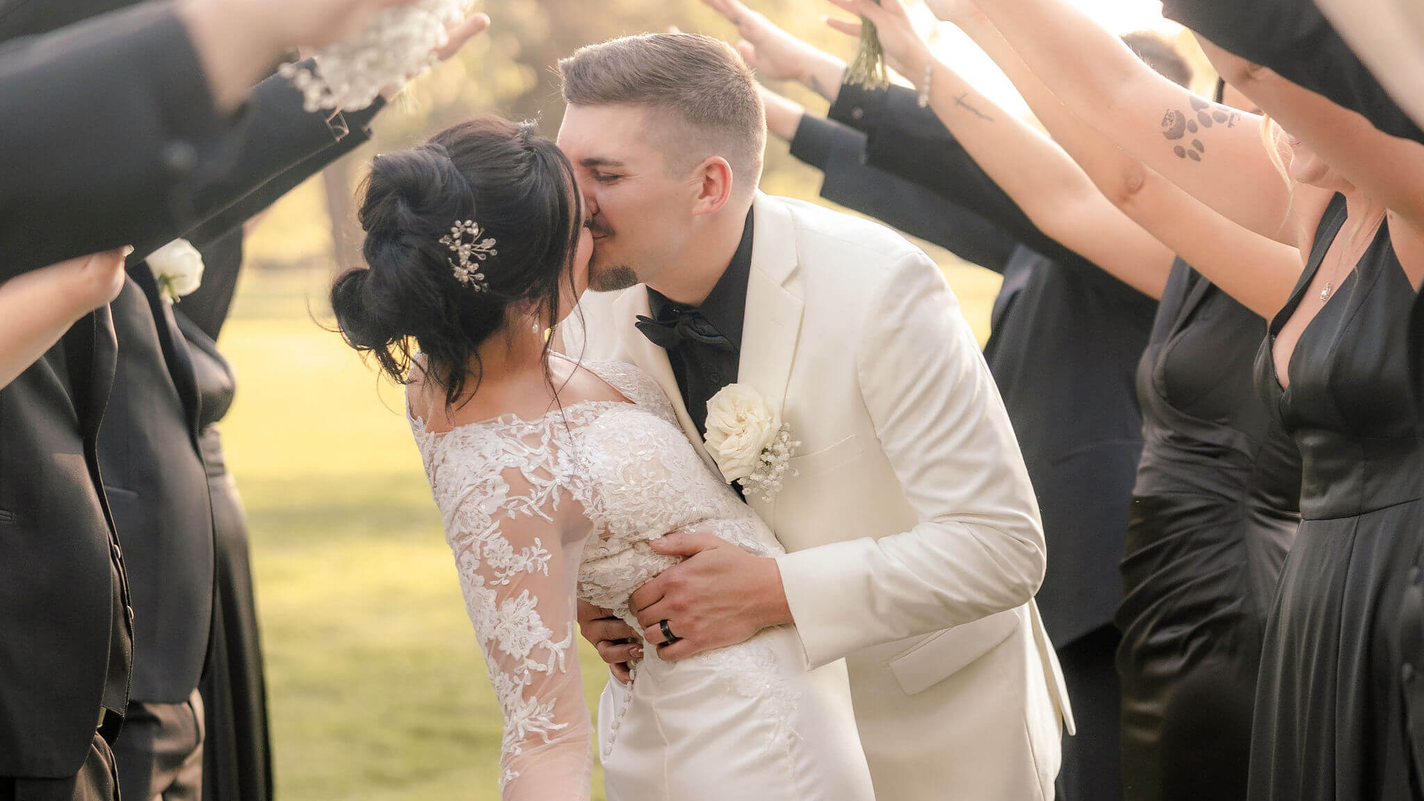 A groom in a white suit dips and kisses the bride, who is wearing a white lace dress, outdoors. People in black outfits stand on each side raising their arms overhead, forming an arch around the couple.