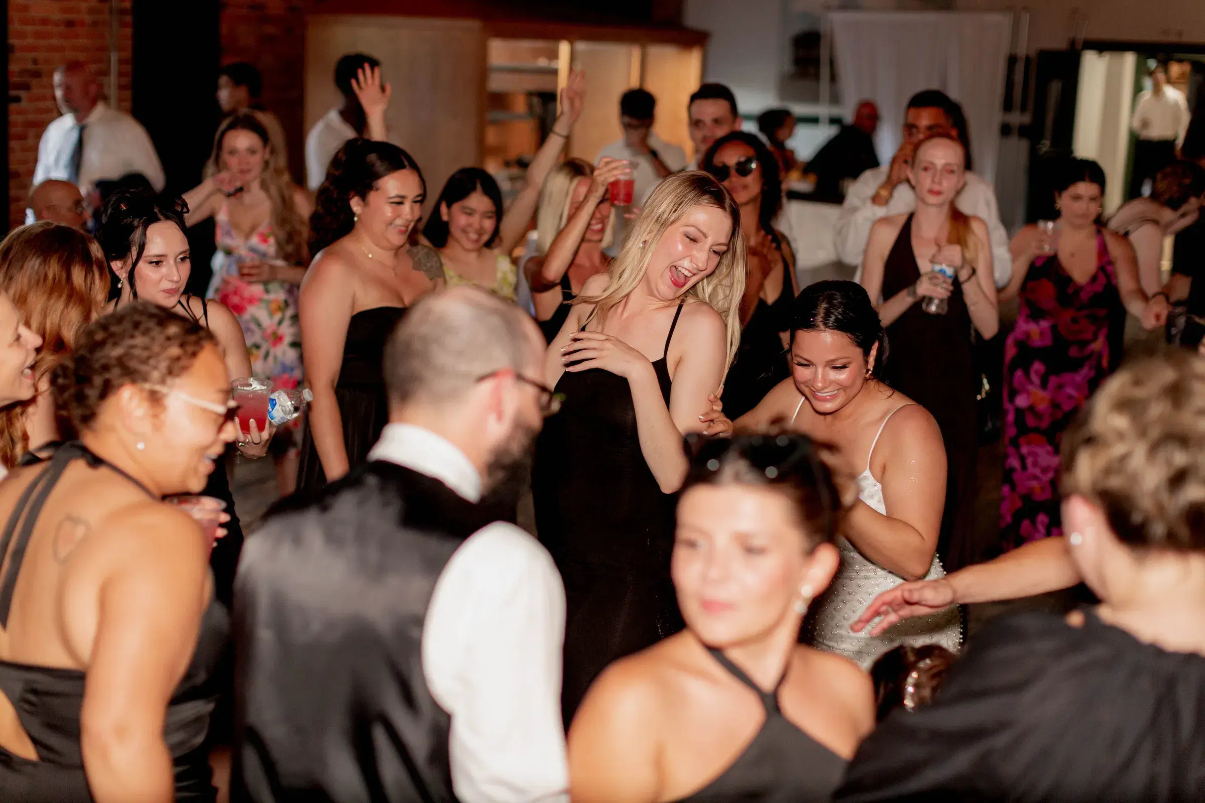 A group of people in formal attire dance and laugh together at an indoor event. Most are wearing black dresses or vests. The setting appears festive, with guests holding drinks and smiling.
