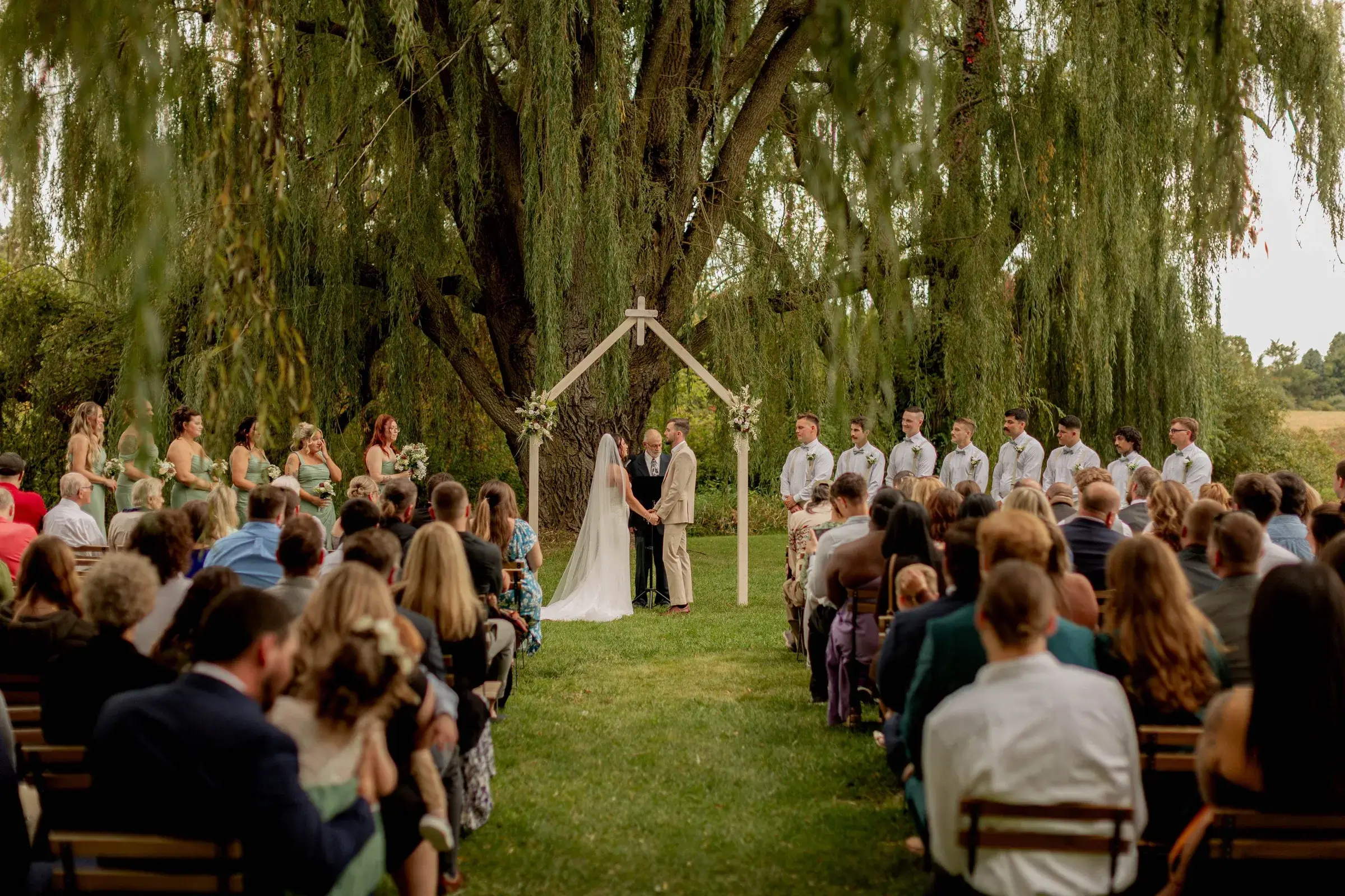 A wedding ceremony taking place outdoors beneath a large tree, with a couple standing under a wooden arch, guests seated on either side, and members of the wedding party standing near the couple.