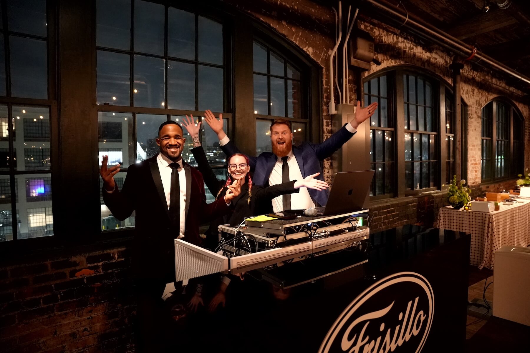 Three people dressed in formal attire stand behind a DJ booth with their arms raised and smiling. The booth is labeled "Frisillo." They are indoors with large windows and exposed brick walls in the background.