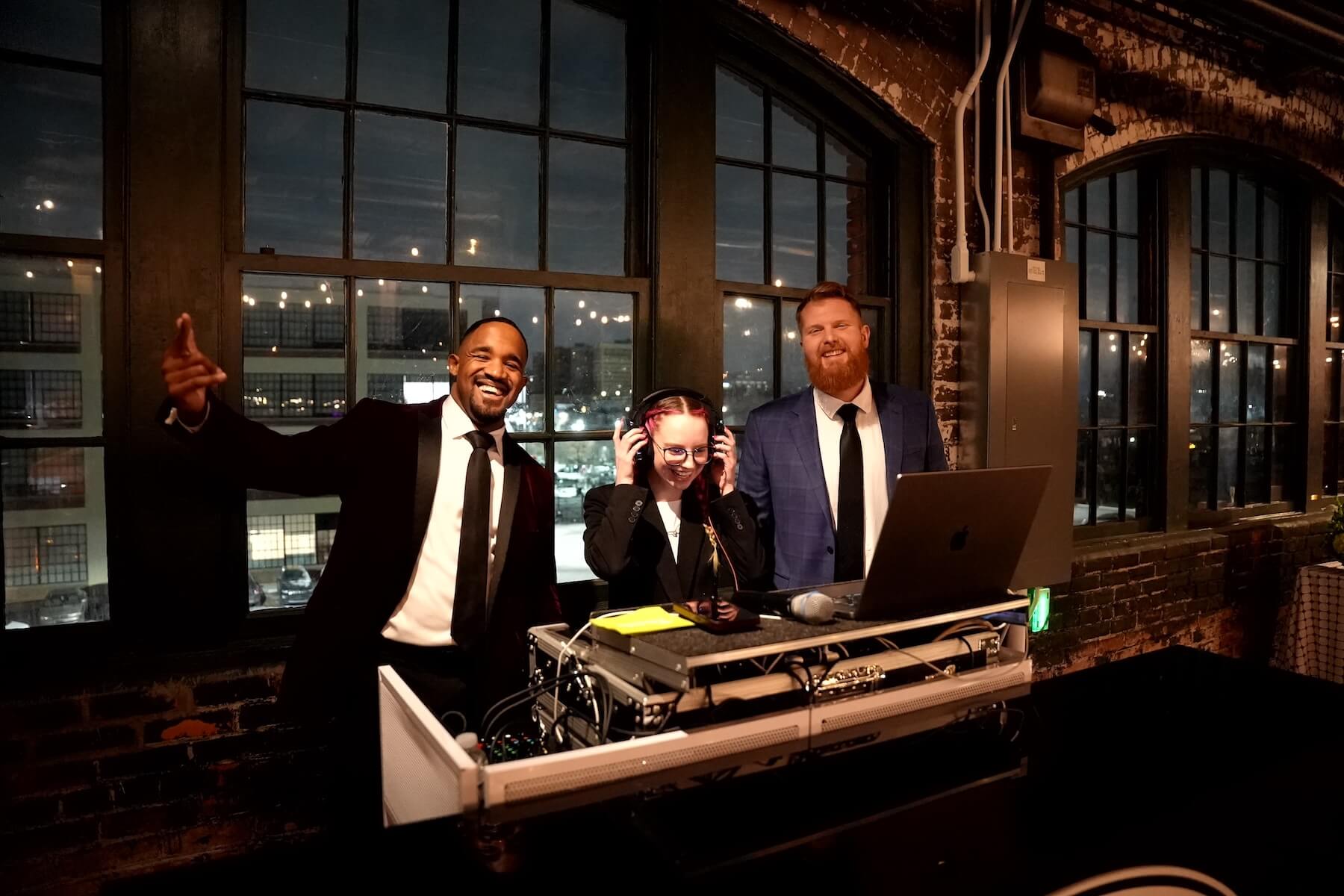 Three people stand behind a DJ booth with equipment. One person in a black suit gives a thumbs up, another wears headphones and smiles, and a third, in a blue suit, stands beside them. Large windows and brick walls are in the background.