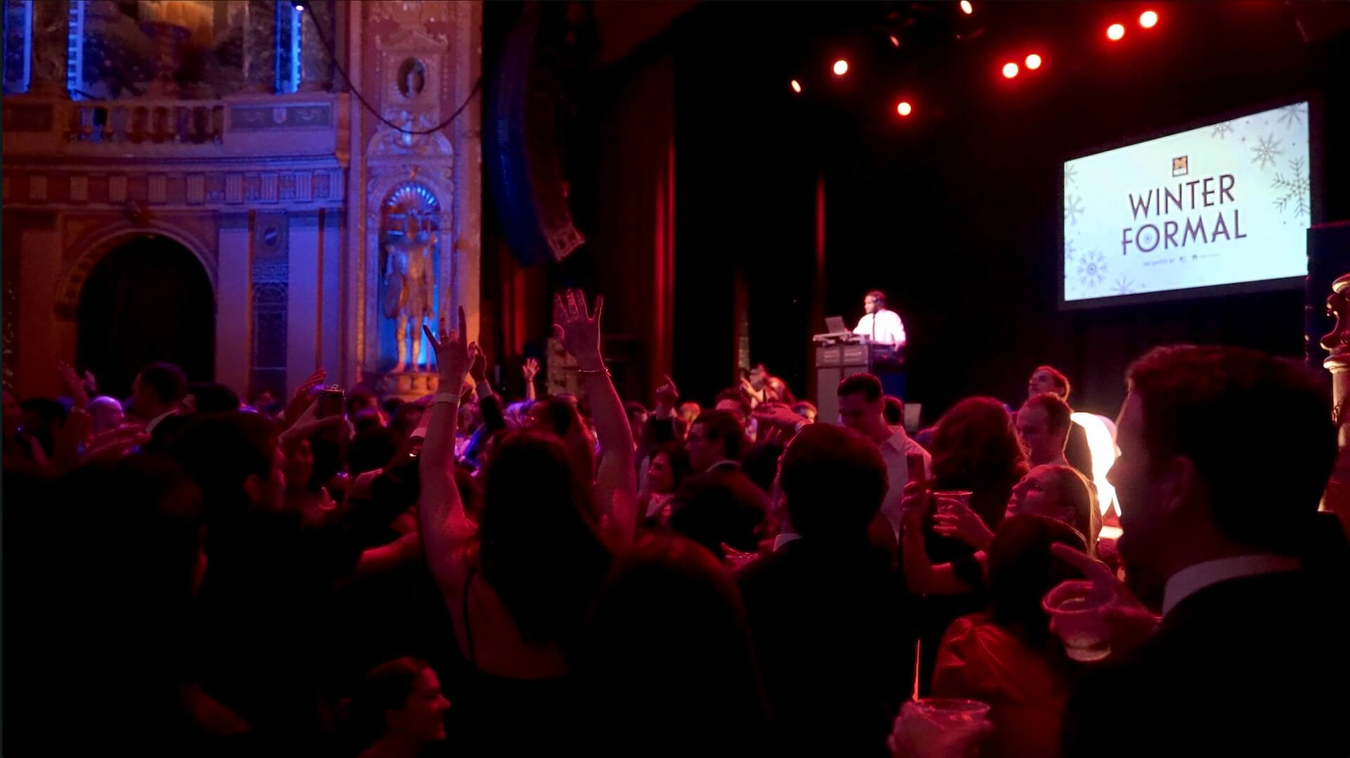 A crowd of people in formal attire stands and raises hands in a decorated theater with red lighting. A person speaks at a podium on stage under a sign that reads "Winter Formal.