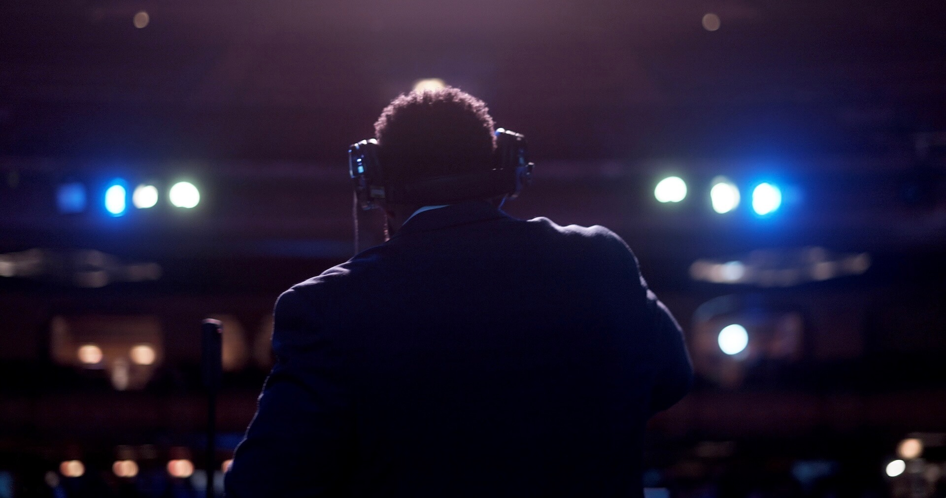 A person wearing headphones and a dark suit stands on a stage facing an audience, with bright stage lights illuminating the background. The image is taken from behind the person.