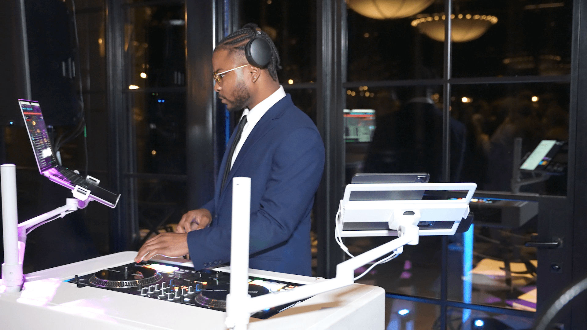A man in a suit wearing headphones stands at a DJ booth, using DJ equipment and a laptop, with another screen mounted nearby. The background shows large windows and dim lighting with reflections.