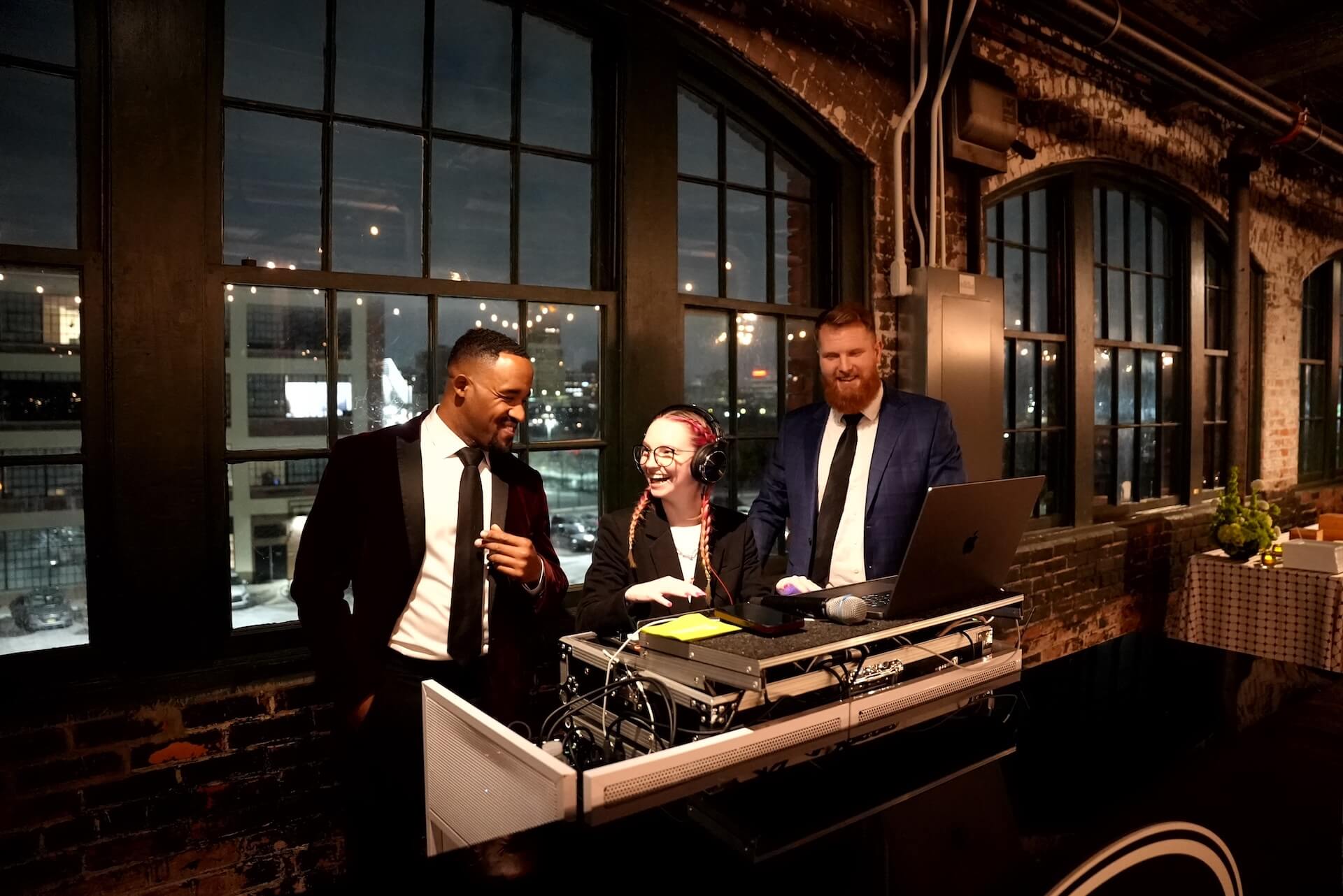 Three people in formal attire stand at a DJ booth inside an industrial-style venue with large windows. One person is at the DJ controls, while the other two stand nearby, all appearing to enjoy the moment.