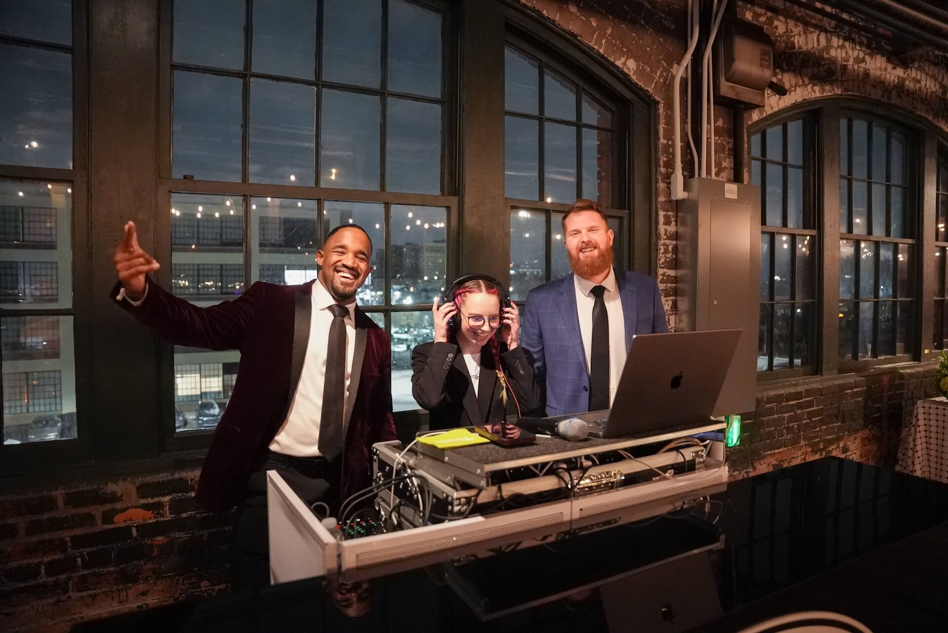 Three people stand behind a DJ booth in a brick-walled room with large windows. One person wears headphones and stands at the DJ equipment, while the other two, both in suits, smile and pose for the photo.