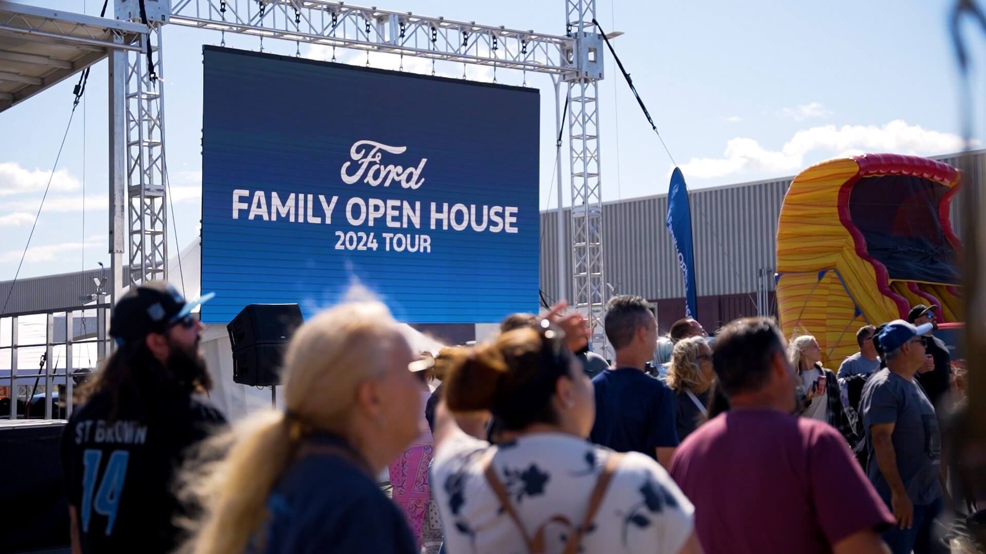 A crowd of people stands outside near a large screen displaying "Ford Family Open House 2024 Tour" at an event; there are stage lights, a blue sky, and part of an inflatable structure in the background.