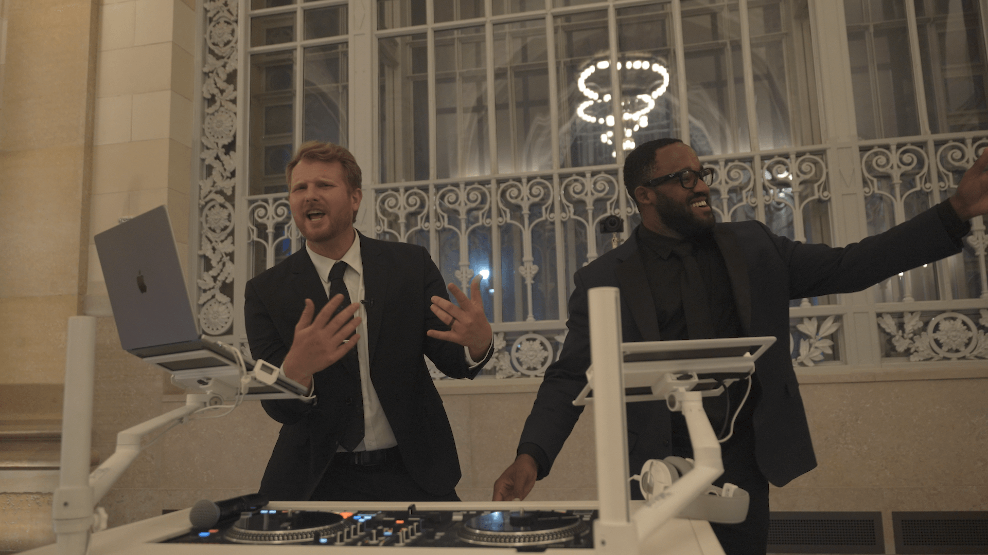Two men in suits stand behind DJ equipment. One gestures expressively while singing or speaking, and the other smiles with an arm raised. They are indoors in front of a large, ornate window.