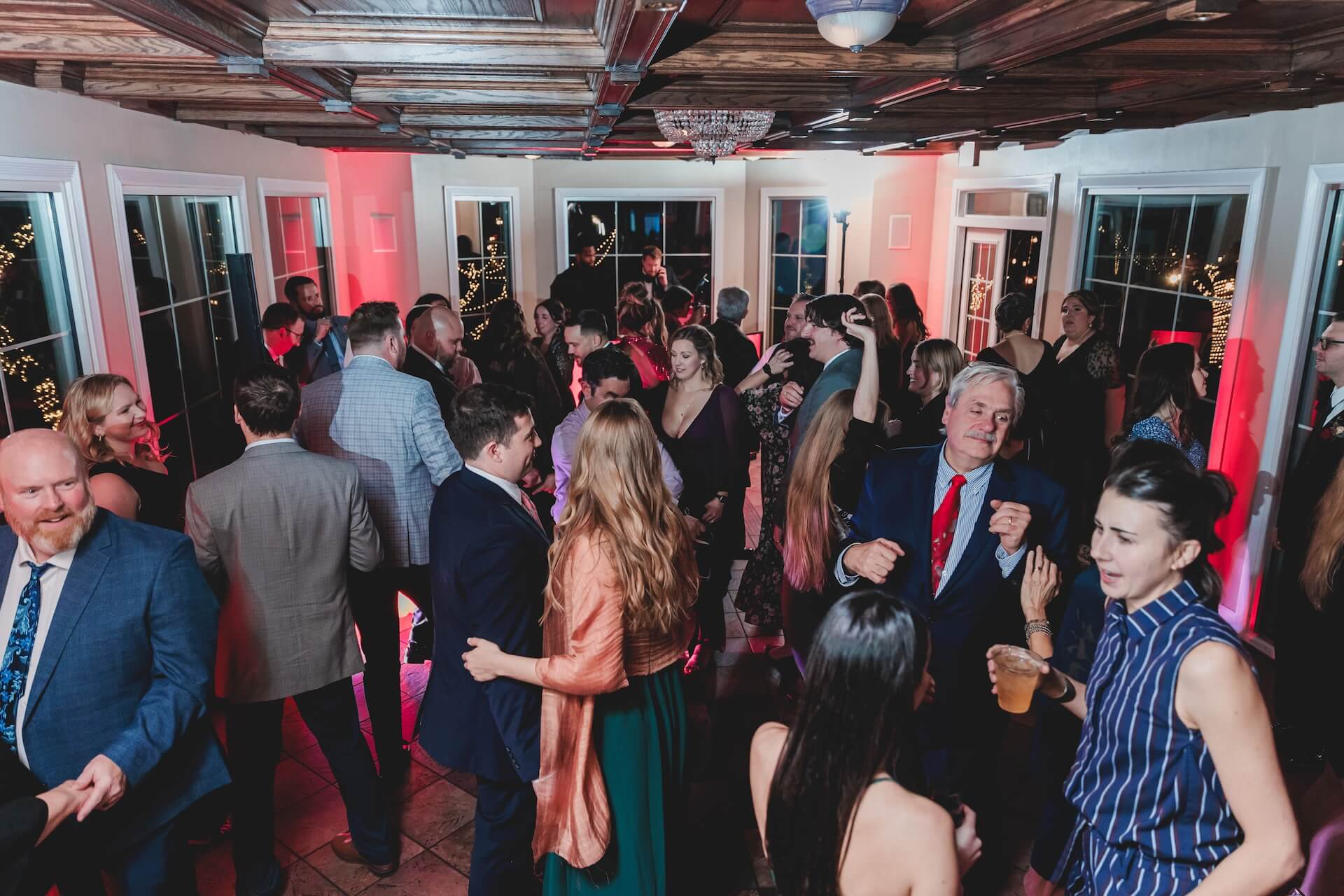 A group of people, dressed in formal and semi-formal attire, are gathered in a well-lit room with large windows and wooden ceiling, socializing and dancing at a party or event.