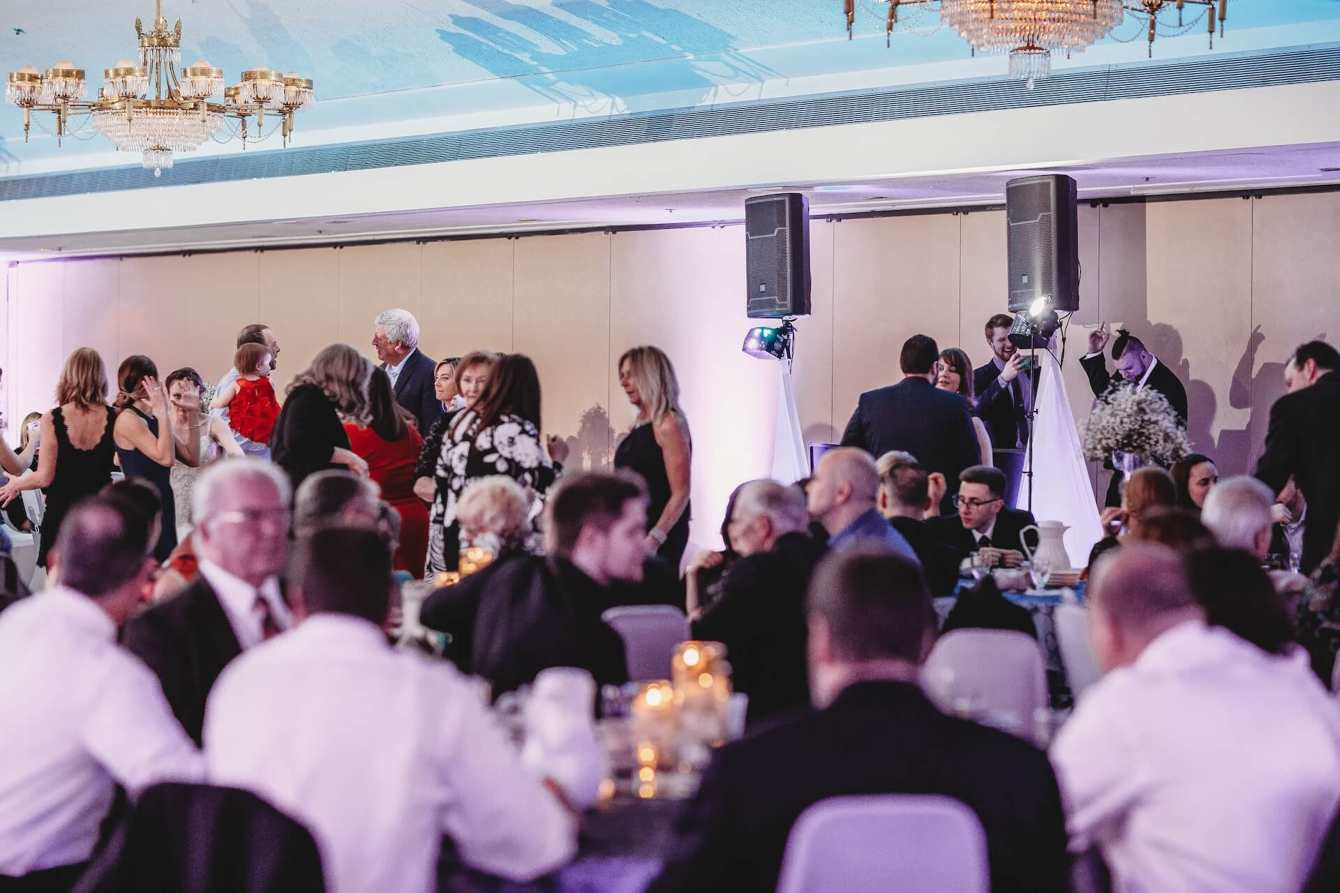 A group of people are seated at round tables with candles, while others stand and talk near a wall in a banquet hall with chandeliers and speakers. The setting appears formal and festive.