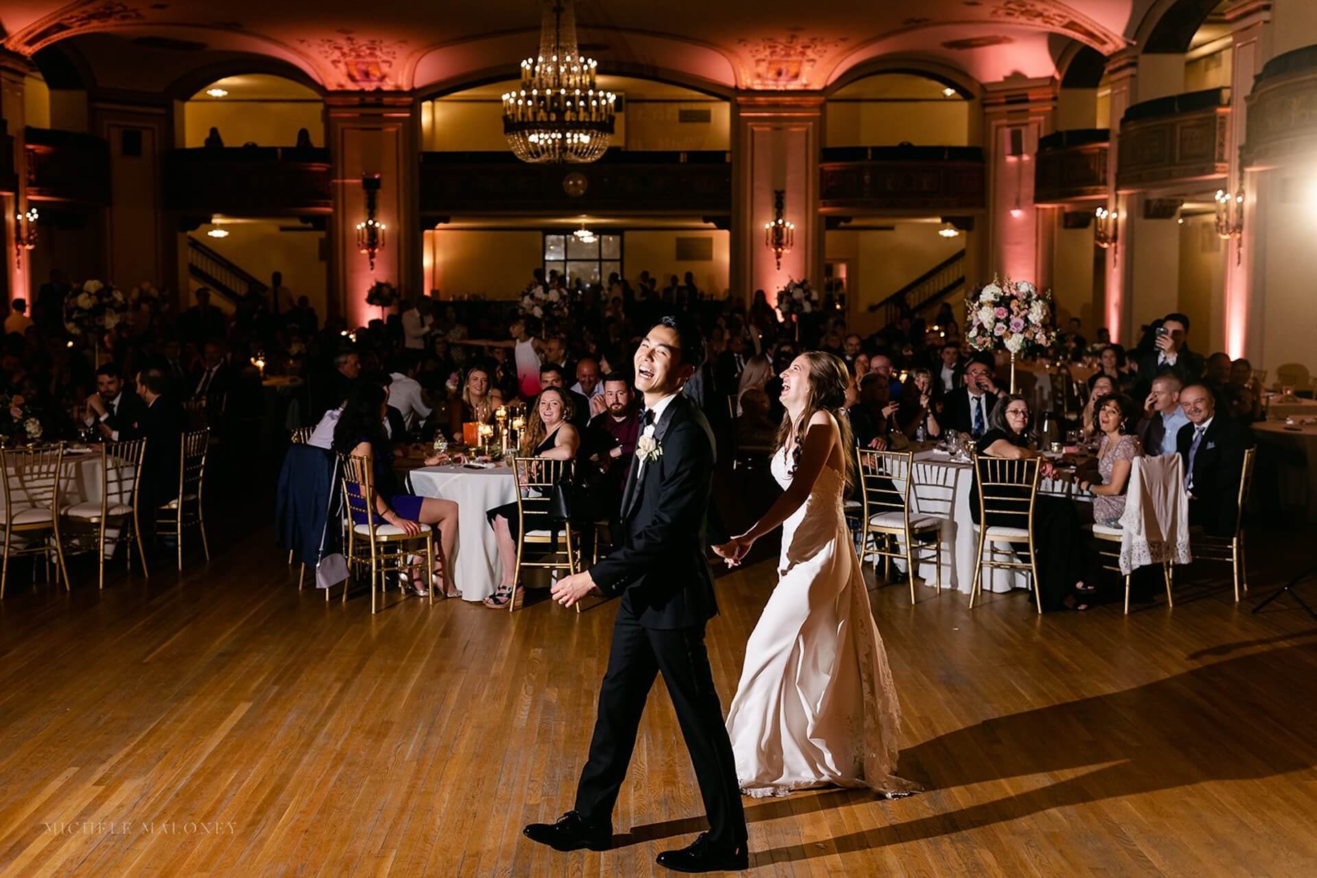 A couple in formal attire holds hands and smiles while walking across a ballroom dance floor, with seated guests watching and tables decorated with flowers in the background. The room is warmly lit with chandeliers and pink uplighting.