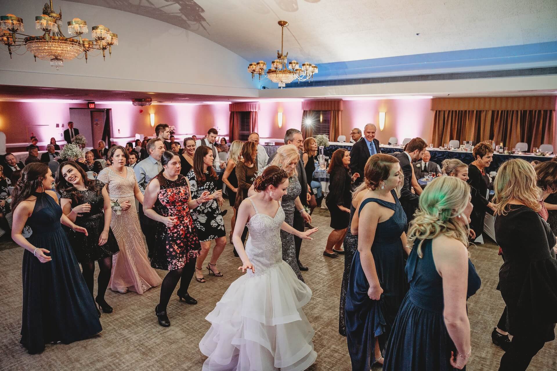 A group of people dressed in formal attire, including a bride in a white gown, dance together in a ballroom with chandeliers and pink uplighting.