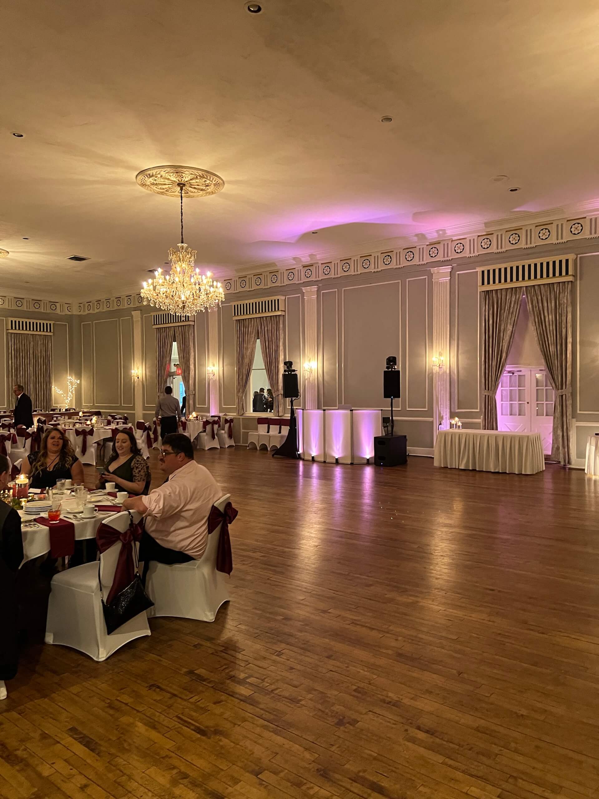 A spacious banquet hall with chandeliers, wood flooring, and tables set for an event. Purple lighting accents the walls, and a small group of people sits at a decorated table on the left side of the image.
