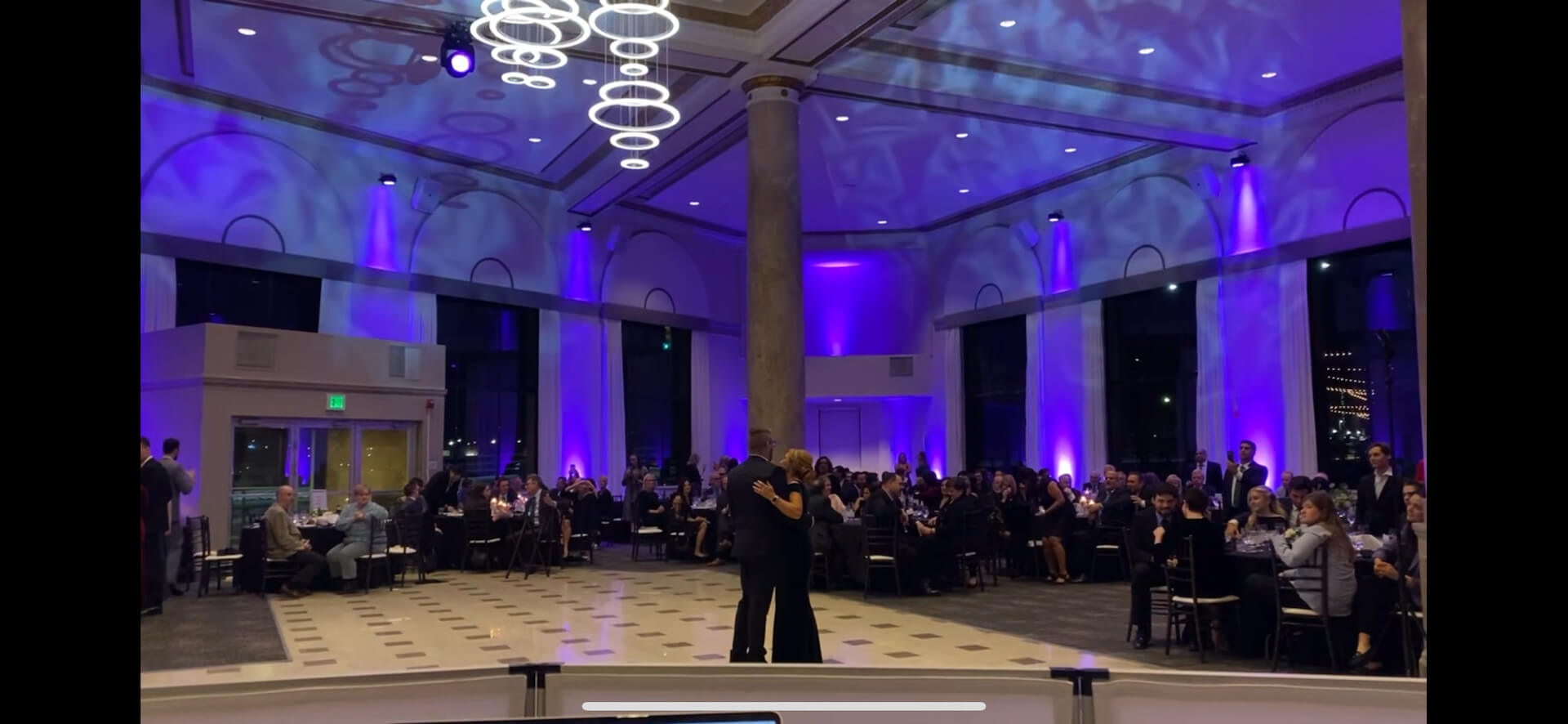 A couple dances in the center of a large, elegant event hall with purple lighting, while guests seated at round tables watch. The ceiling is high with modern circular chandeliers.