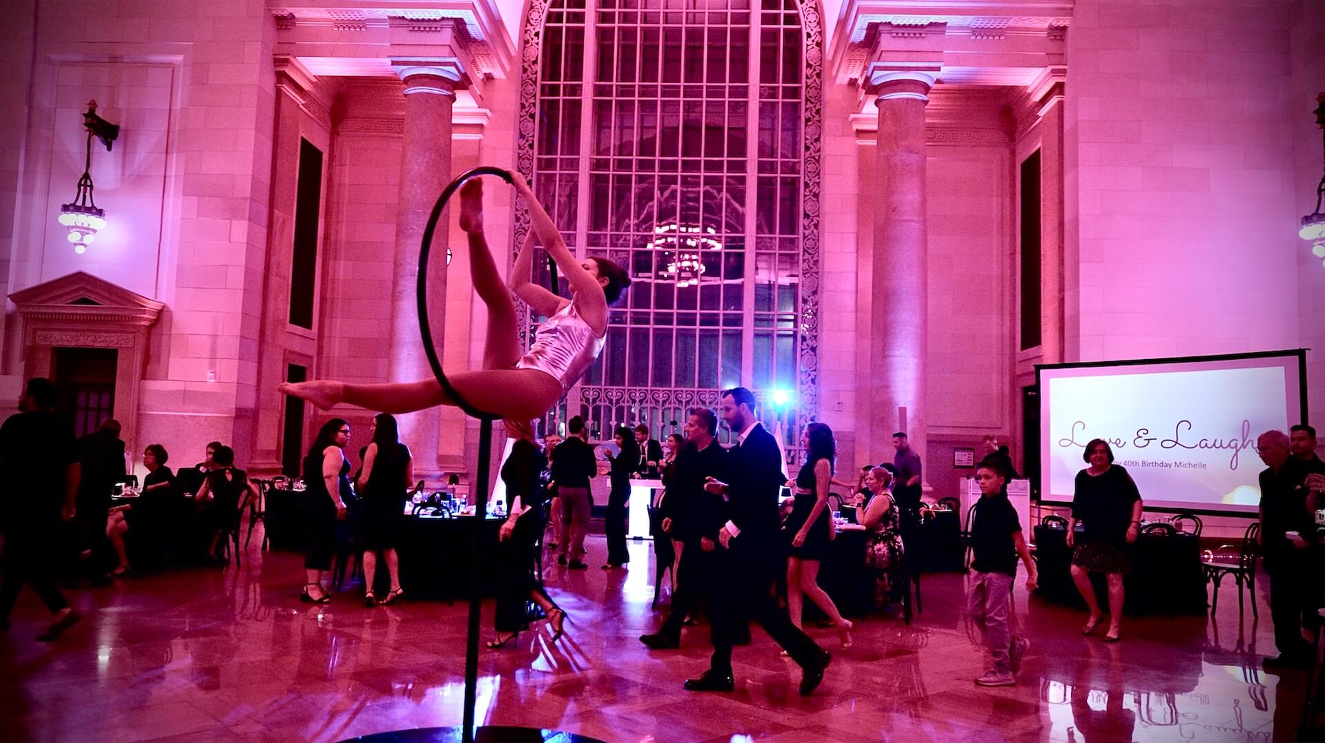 A performer in a silver costume poses on an aerial hoop in the center of a large, ornate venue with high ceilings and purple lighting. Guests in formal attire watch and mingle around tables in the background.