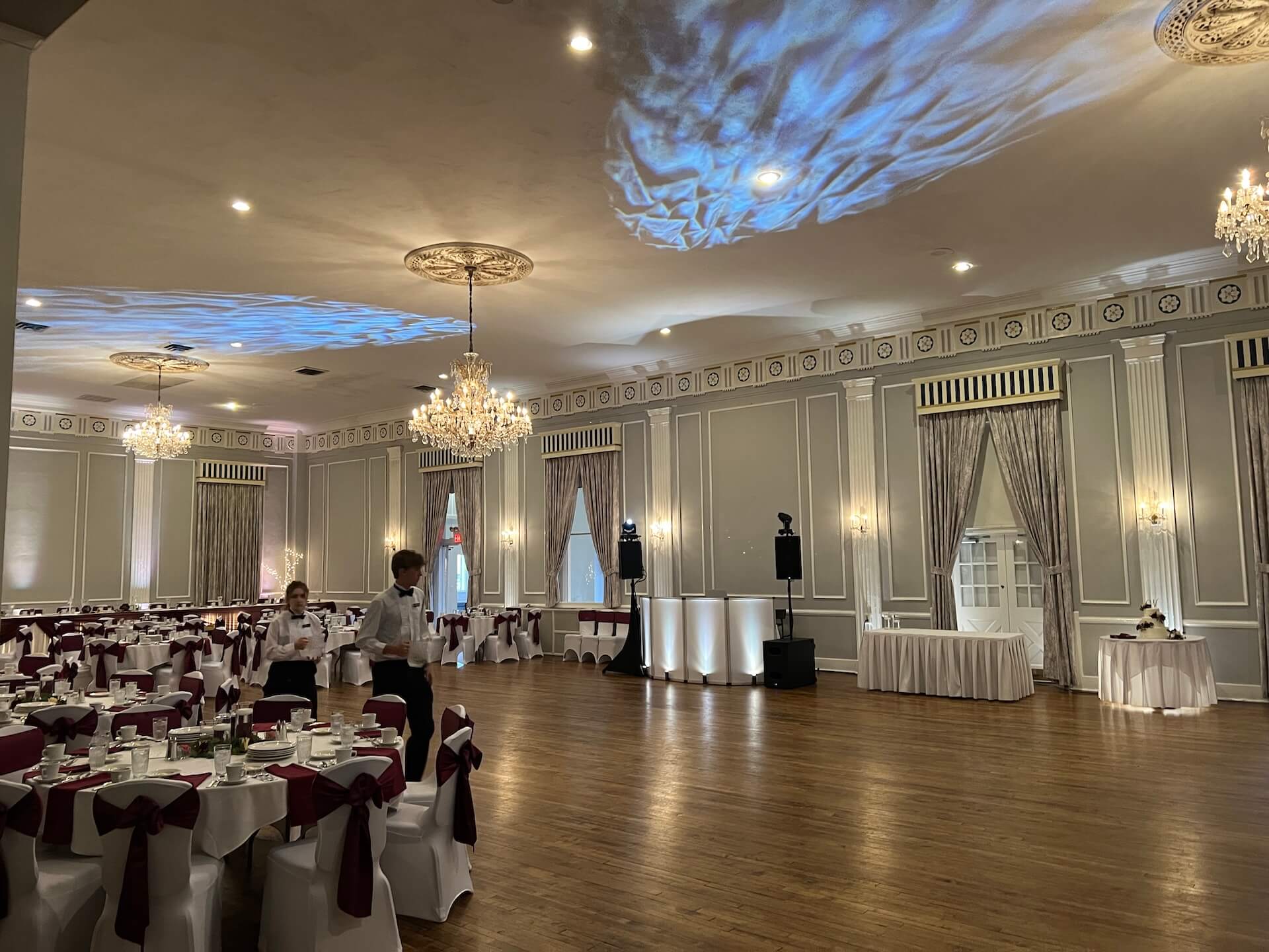 Large banquet hall set for an event, with round tables covered in white tablecloths and burgundy chair sashes, chandeliers on the ceiling, and staff members walking among the tables. A DJ booth is set up near the dance floor.