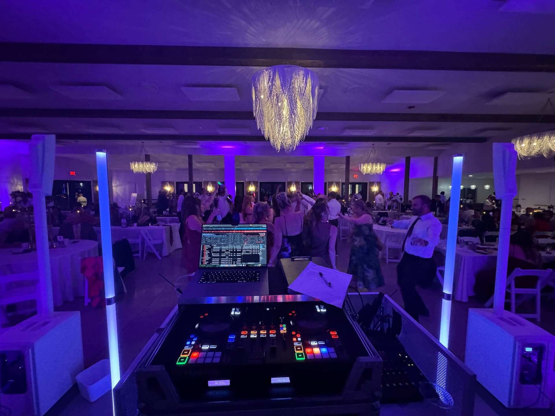 A view from behind a DJ booth with illuminated equipment, facing a crowded dance floor at an indoor event under purple lighting and decorative chandeliers. People are dancing and tables are visible in the background.