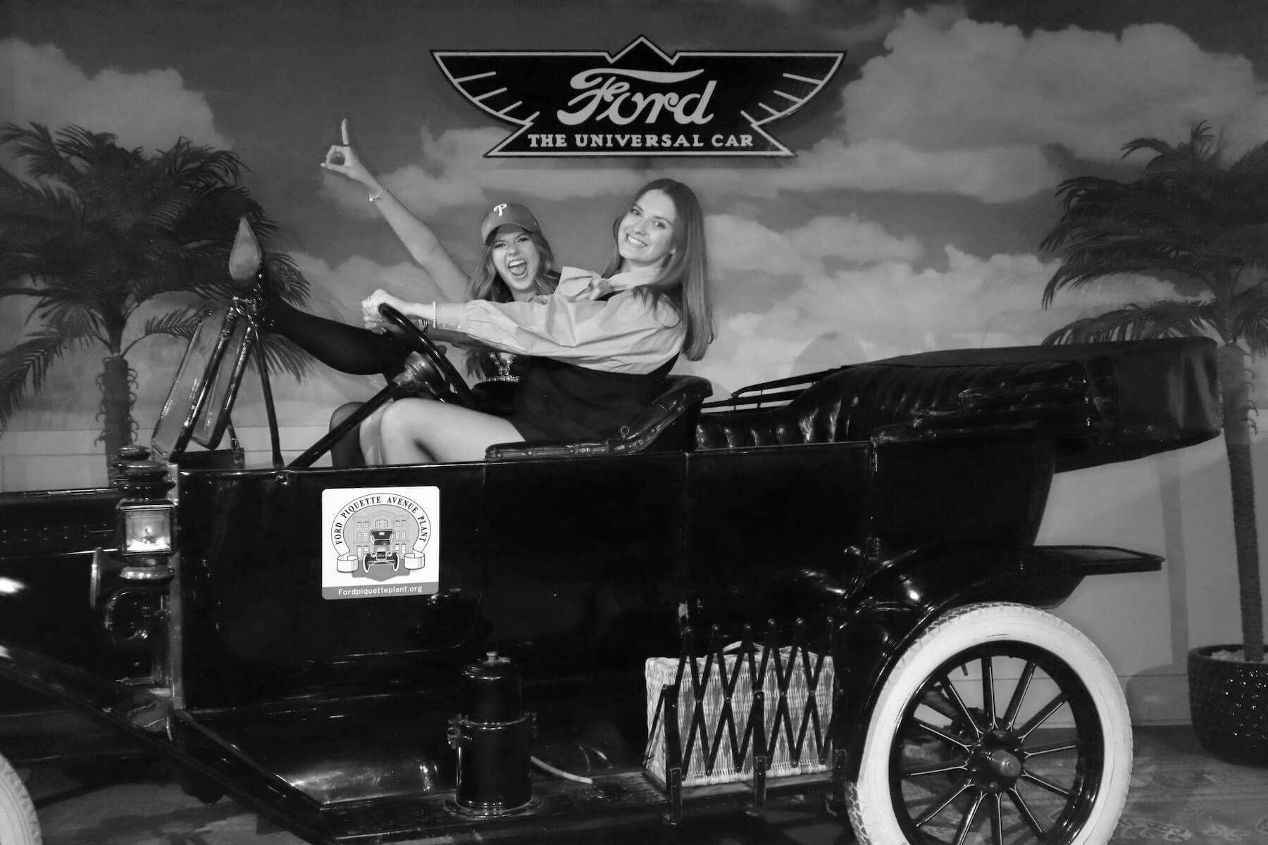 Two women pose and smile in an old-fashioned Ford car display, with "Ford The Universal Car" sign above them. Both appear to be enjoying themselves. The scene is set against a backdrop with palm trees and clouds.
