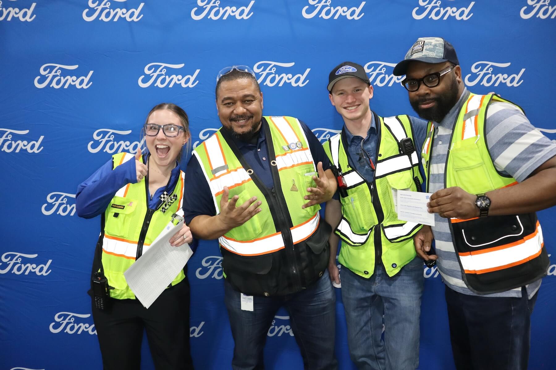 Four people wearing safety vests and holding papers stand smiling in front of a blue Ford-branded backdrop. They appear to be posing for a group photo, with some making hand gestures.