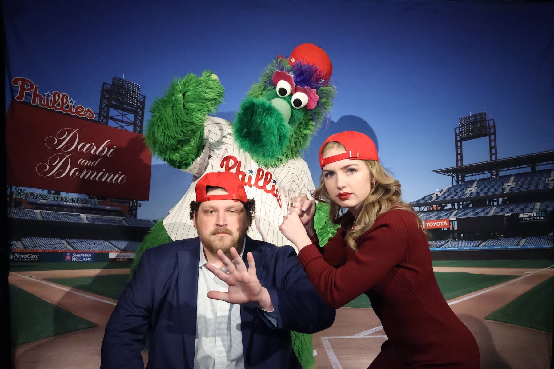 A man in a suit and a woman in a red dress pose seriously at a baseball stadium backdrop with the Phillie Phanatic mascot behind them. Both wear red baseball caps. The mascot raises a fist in the background.