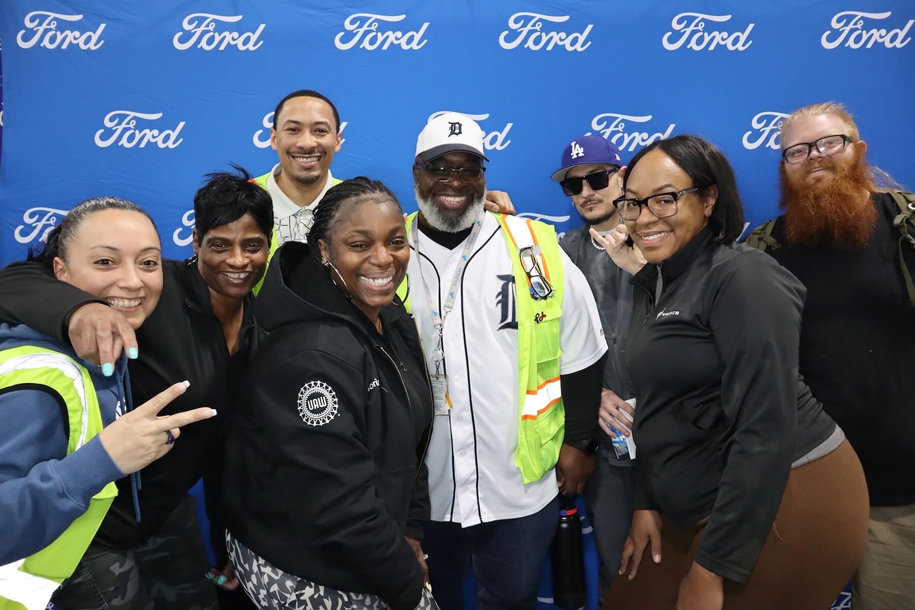 A group of eight people stand smiling in front of a blue Ford backdrop. Most are wearing casual clothes or safety vests, and one person wears a Detroit Tigers jersey and hat.