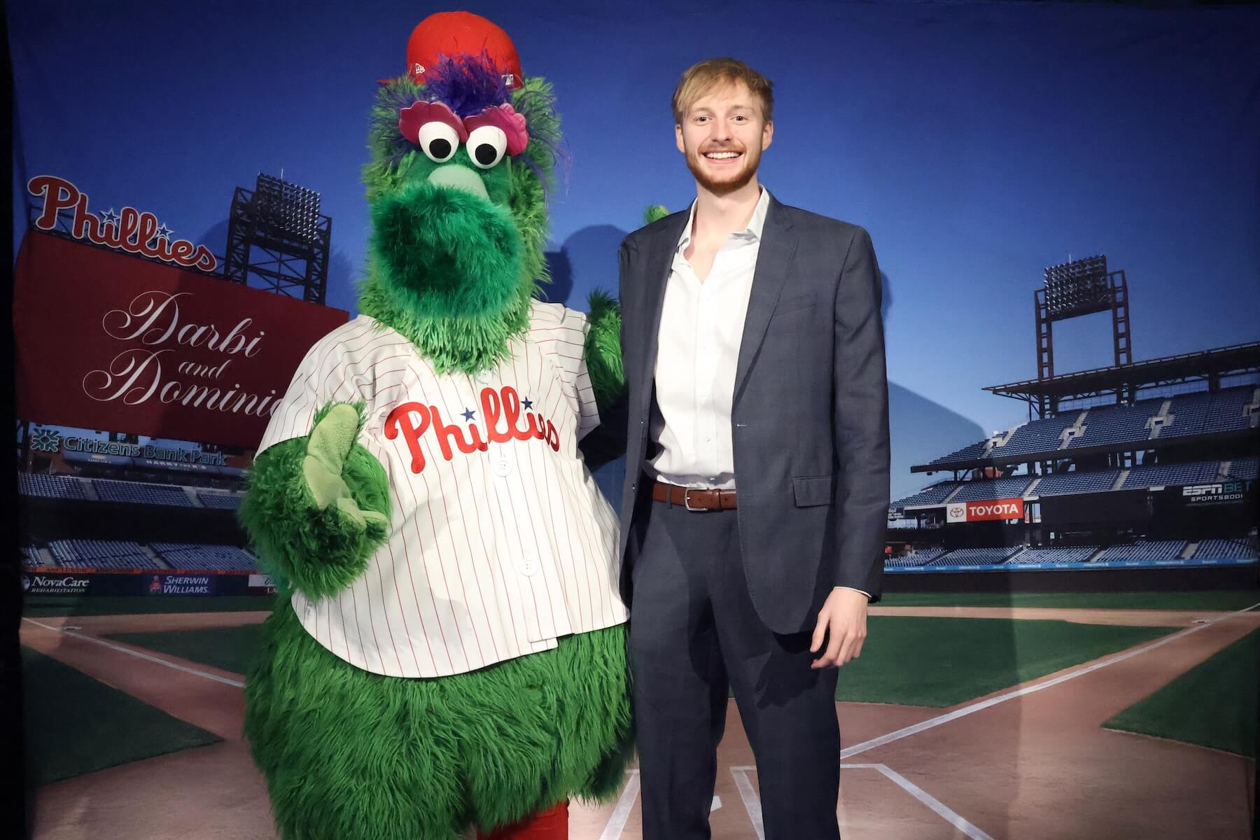 A man in a suit stands next to the Phillie Phanatic mascot on a baseball field with empty stadium seating in the background. Both are smiling at the camera.