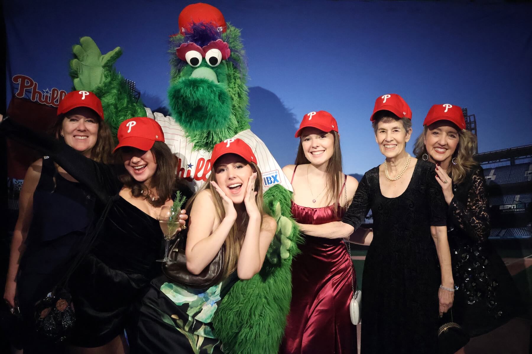 Six women wearing red Phillies baseball caps pose for a photo with the Phillie Phanatic mascot in front of a stadium backdrop, all smiling and appearing to enjoy the moment.