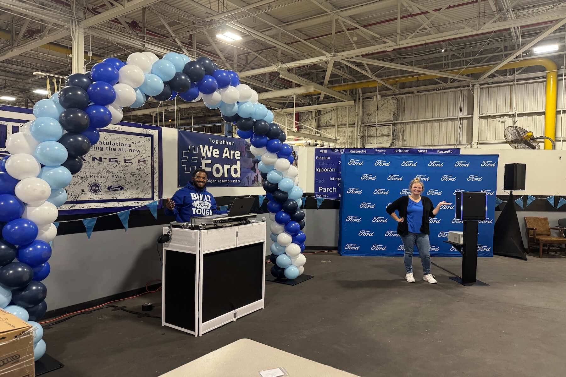 A person stands at a DJ booth under a blue and white balloon arch, while another person in blue speaks at a podium. Ford logos and signs are displayed in a large industrial indoor space.