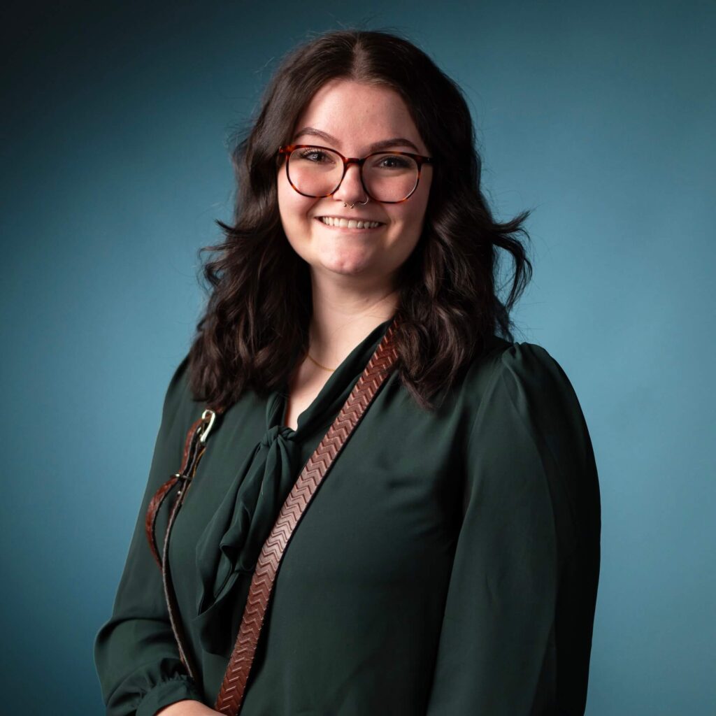 A woman with wavy dark hair and glasses stands in front of a blue background. She is wearing a dark green blouse and a brown strap across her shoulder, smiling at the camera.