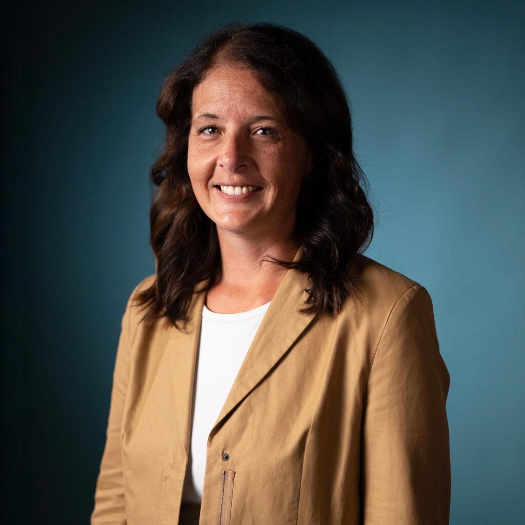A woman with shoulder-length wavy brown hair is wearing a tan blazer over a white top. She is smiling and standing in front of a plain blue background.