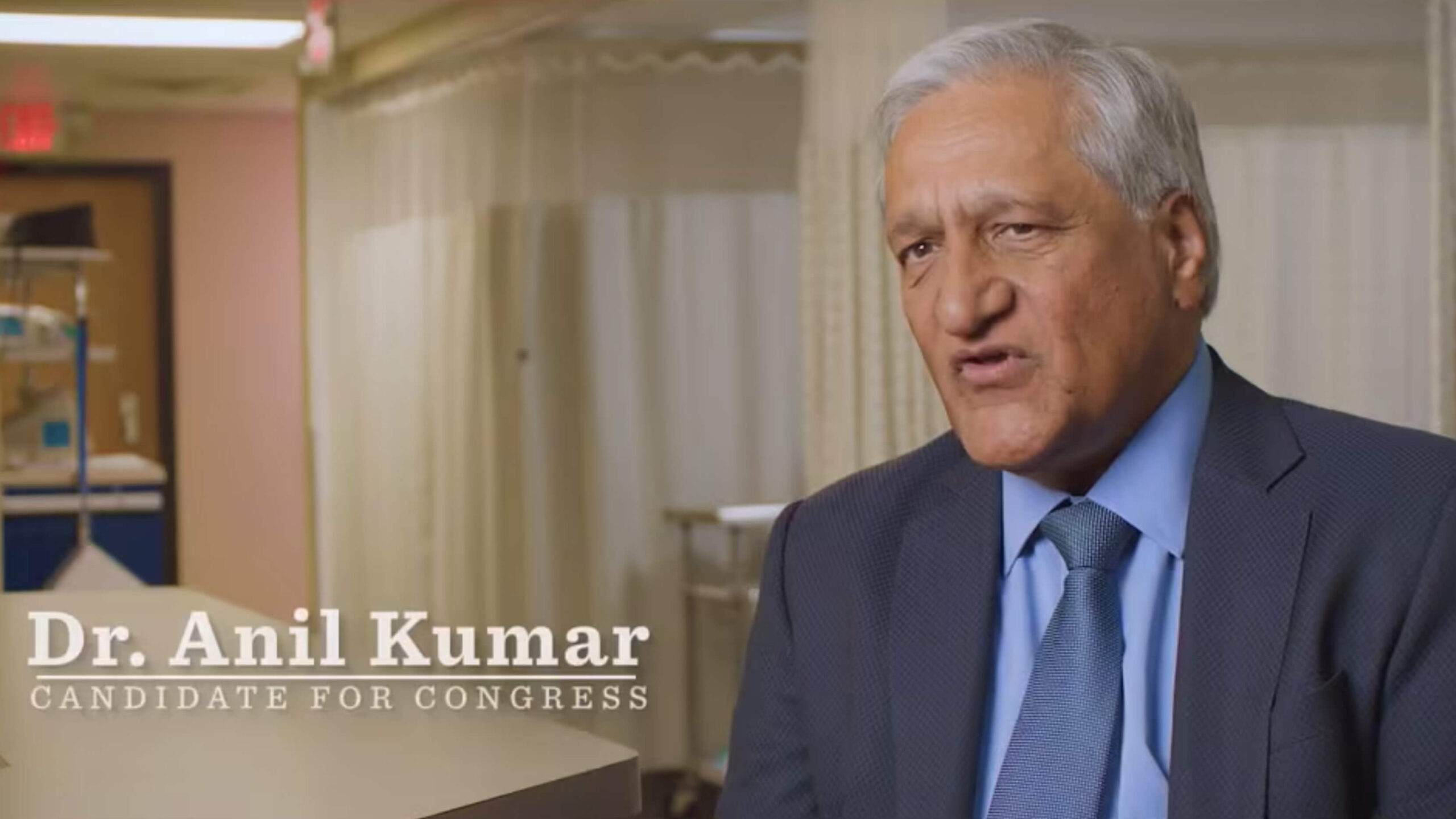 Dr. Anil Kumar, wearing a blue suit and tie, sits indoors and speaks. Text on the image reads, "Dr. Anil Kumar, Candidate for Congress." The background shows curtains and medical equipment.