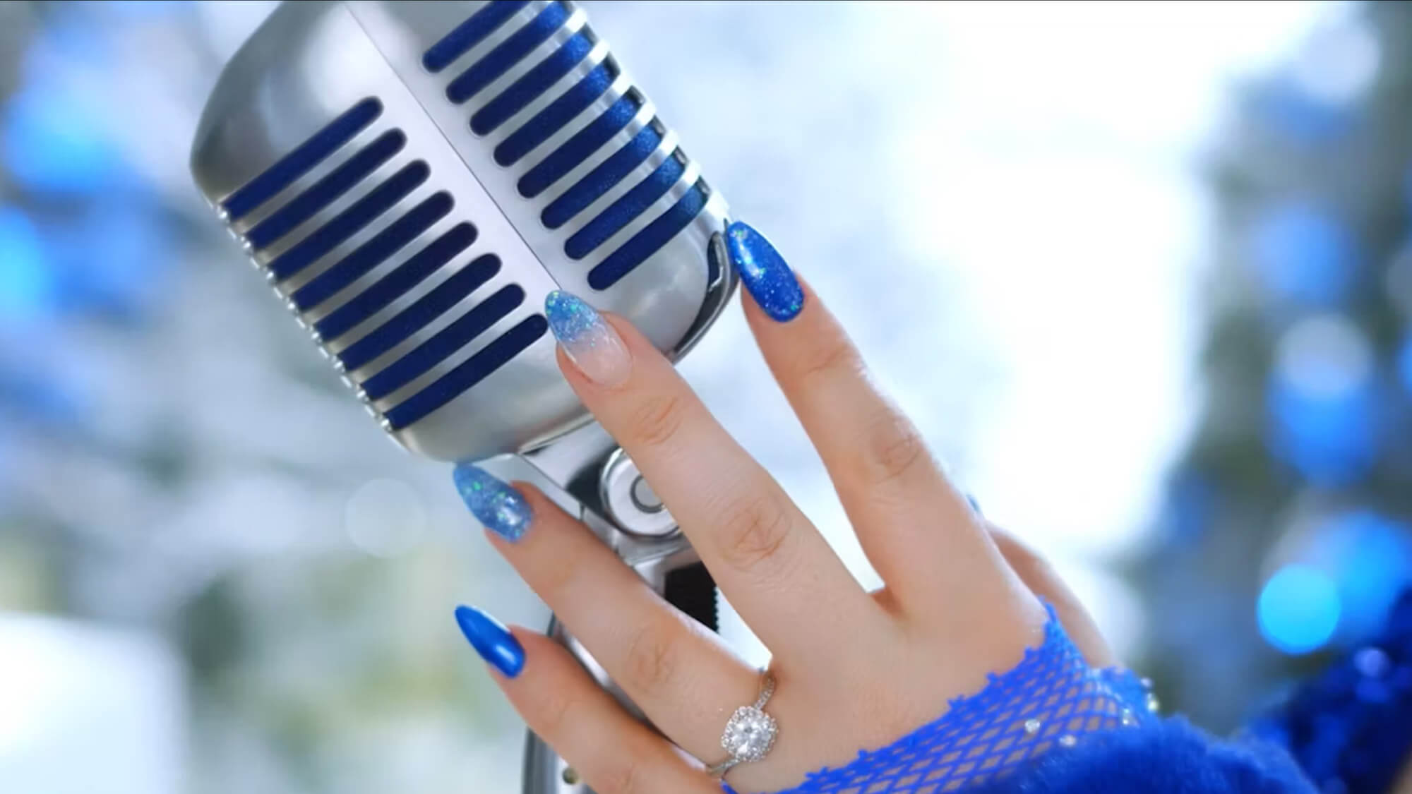 A hand with blue and glittery acrylic nails holds a vintage-style silver microphone. The person is wearing a silver ring with a large round gemstone and a blue fishnet sleeve. The background is blurred with blue and white tones.