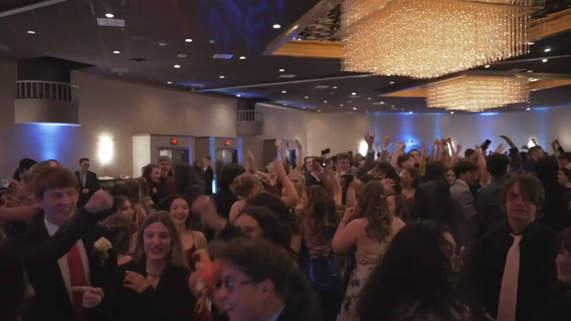 A large group of people dressed in formal attire are dancing and celebrating in a ballroom with chandeliers overhead. Many have their hands raised, and the atmosphere appears lively and festive.