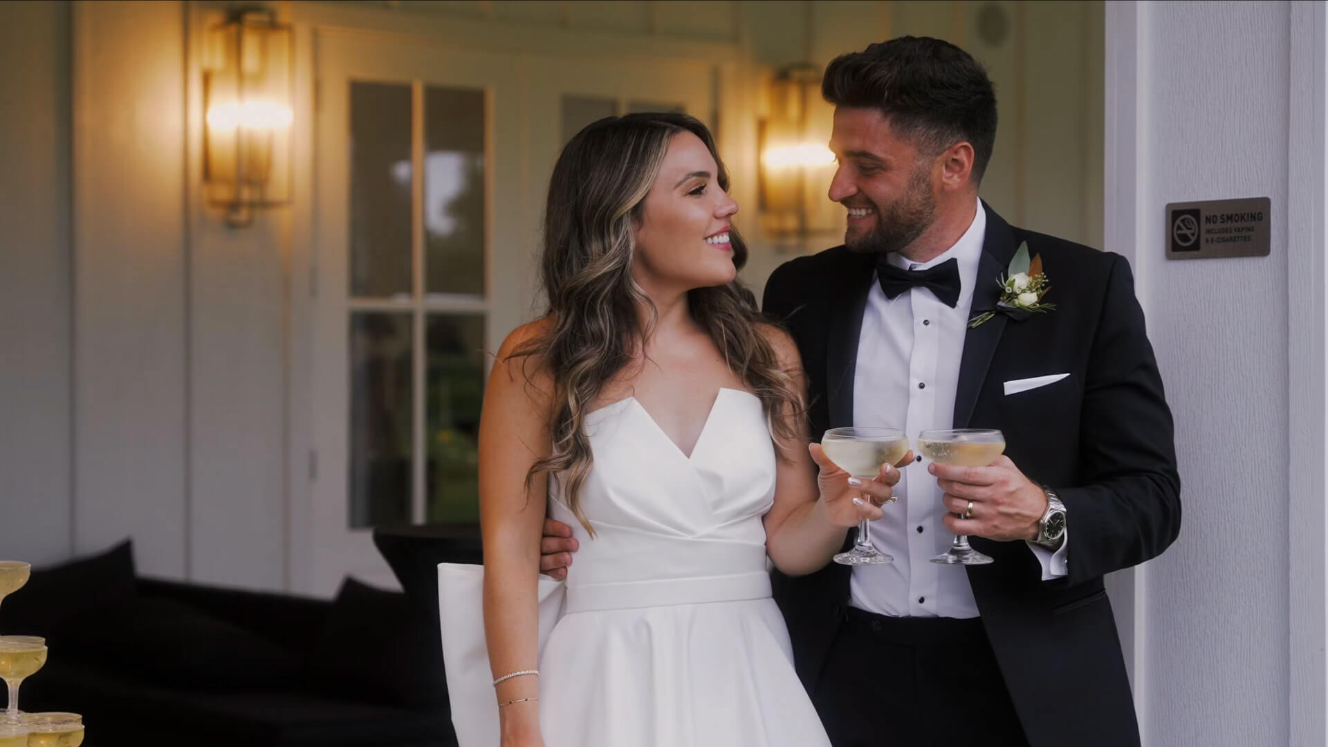 A bride in a white dress and a groom in a black tuxedo smile at each other while holding champagne glasses, standing outside a building with light fixtures and glass doors in the background.