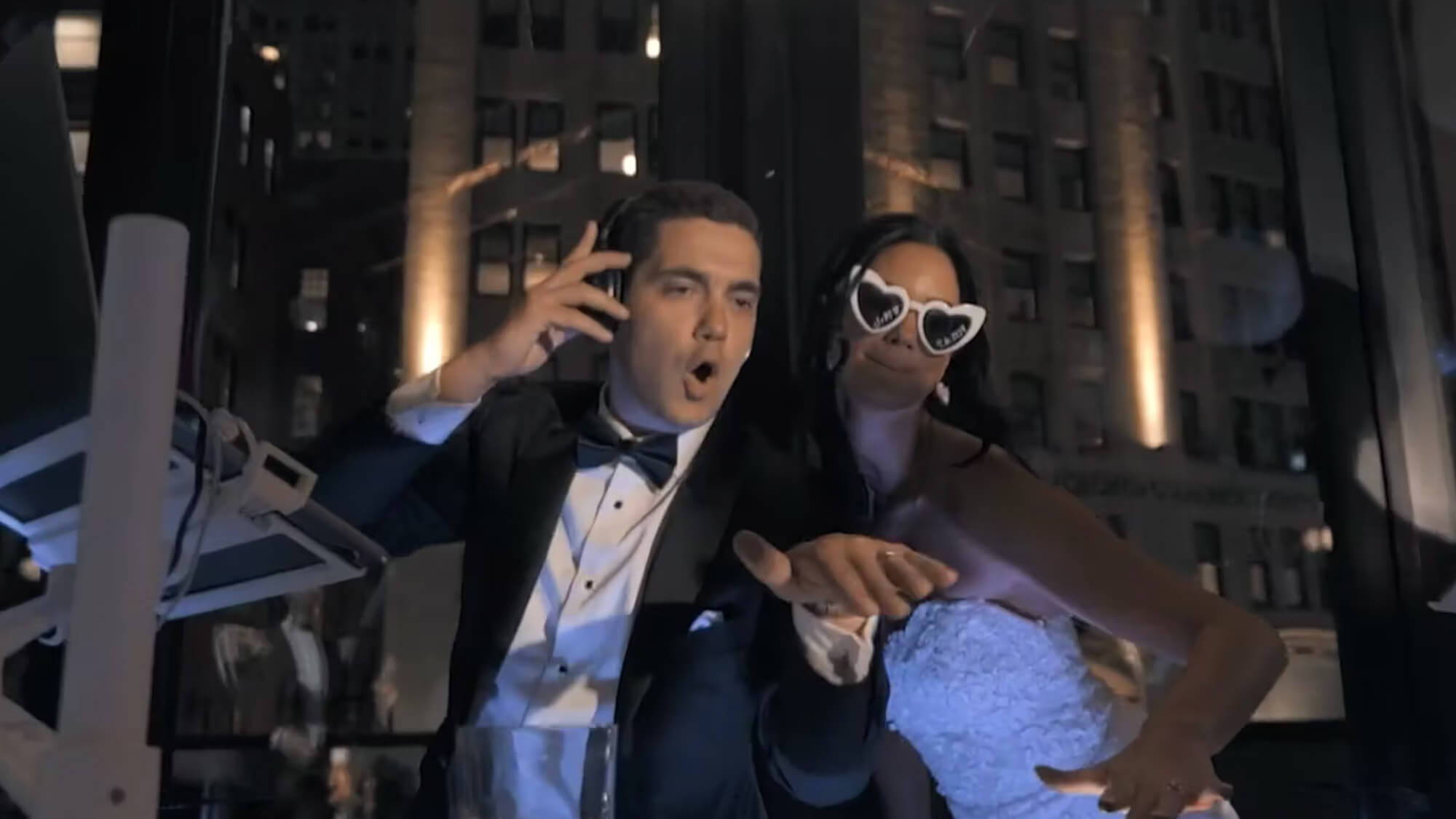 A man in a tuxedo and a woman in a white dress with heart-shaped sunglasses pose together at night indoors; the man is holding a phone to his ear and making an animated expression, while the woman gestures playfully.