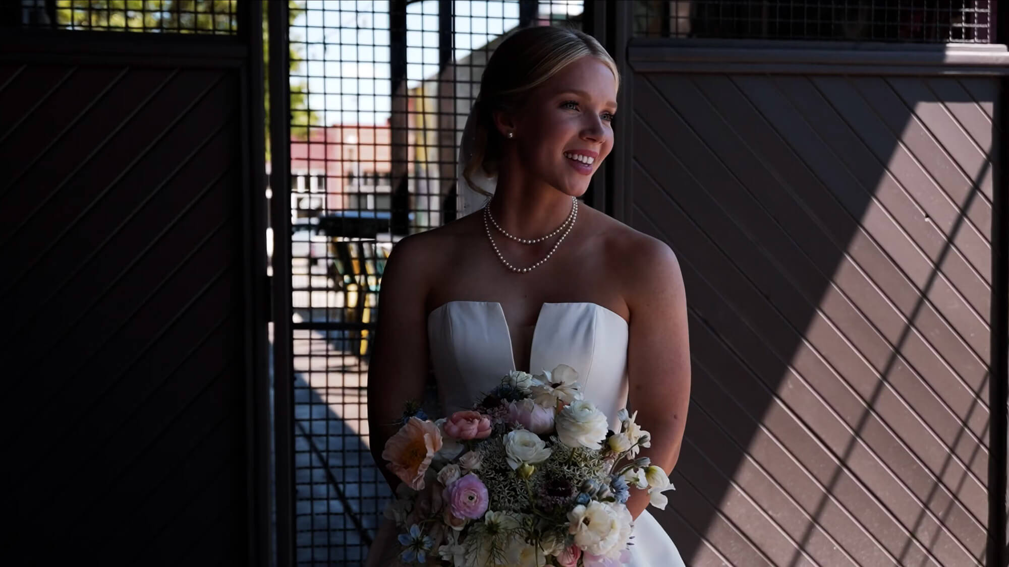 A bride in a strapless white gown and pearl necklace holds a bouquet of flowers while standing indoors in front of a large open doorway with metal bars. Sunlight partially illuminates her and the background.