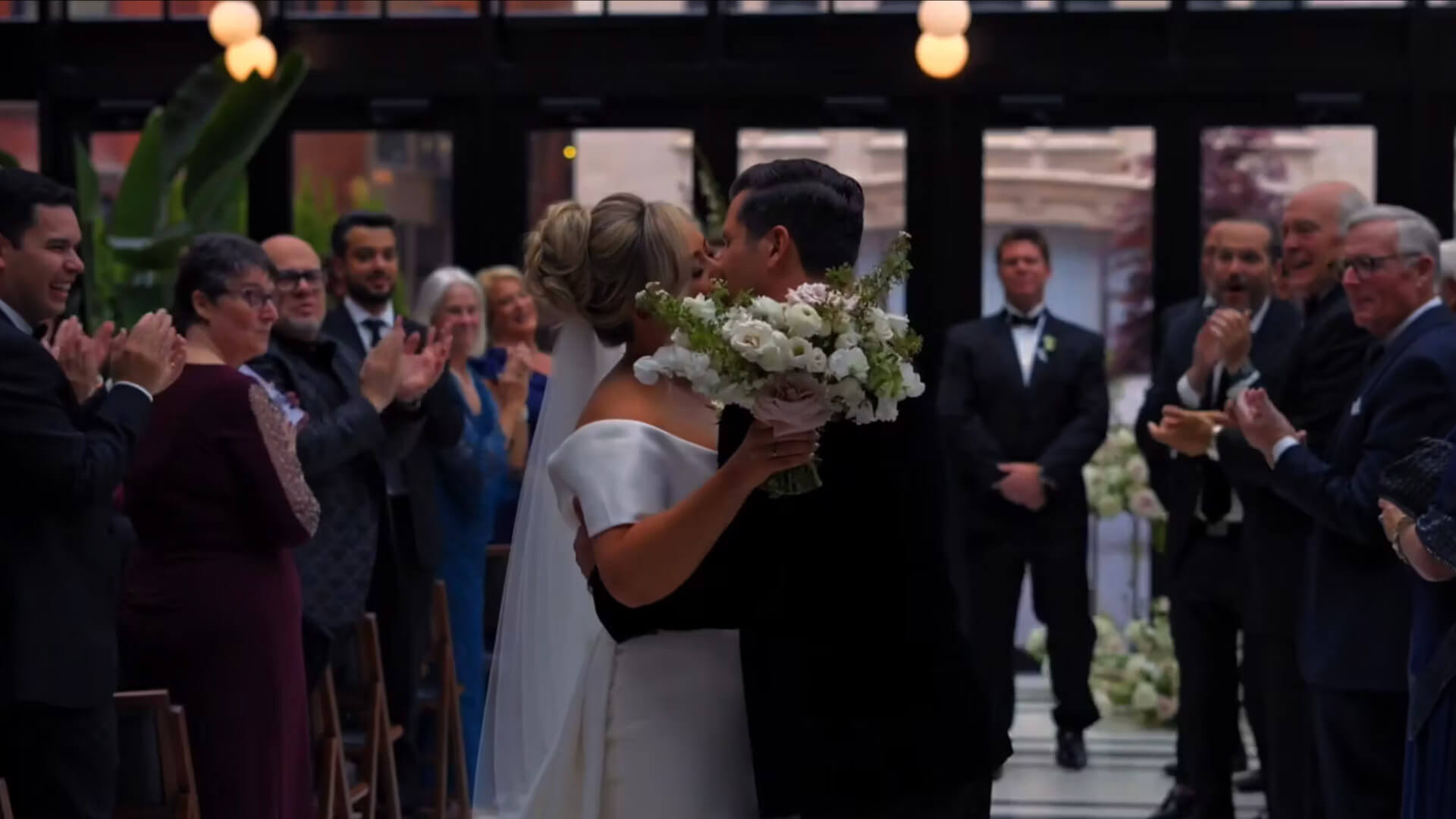 A bride and groom kiss at the end of a wedding aisle as guests on both sides stand and applaud. The bride holds a bouquet of white flowers, and both wear formal wedding attire.